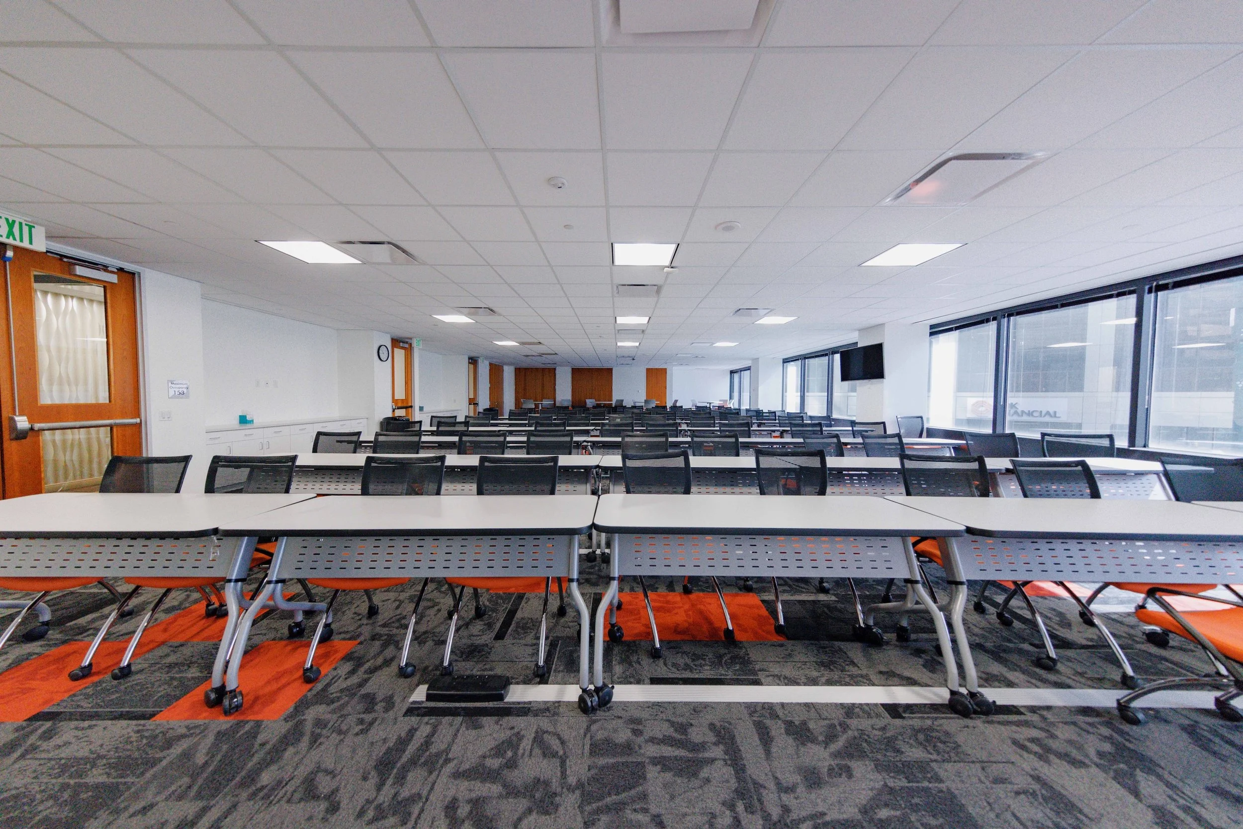 Empty conference room with rows of tables and chairs, large windows on the right, and a flat-screen TV on the right wall.