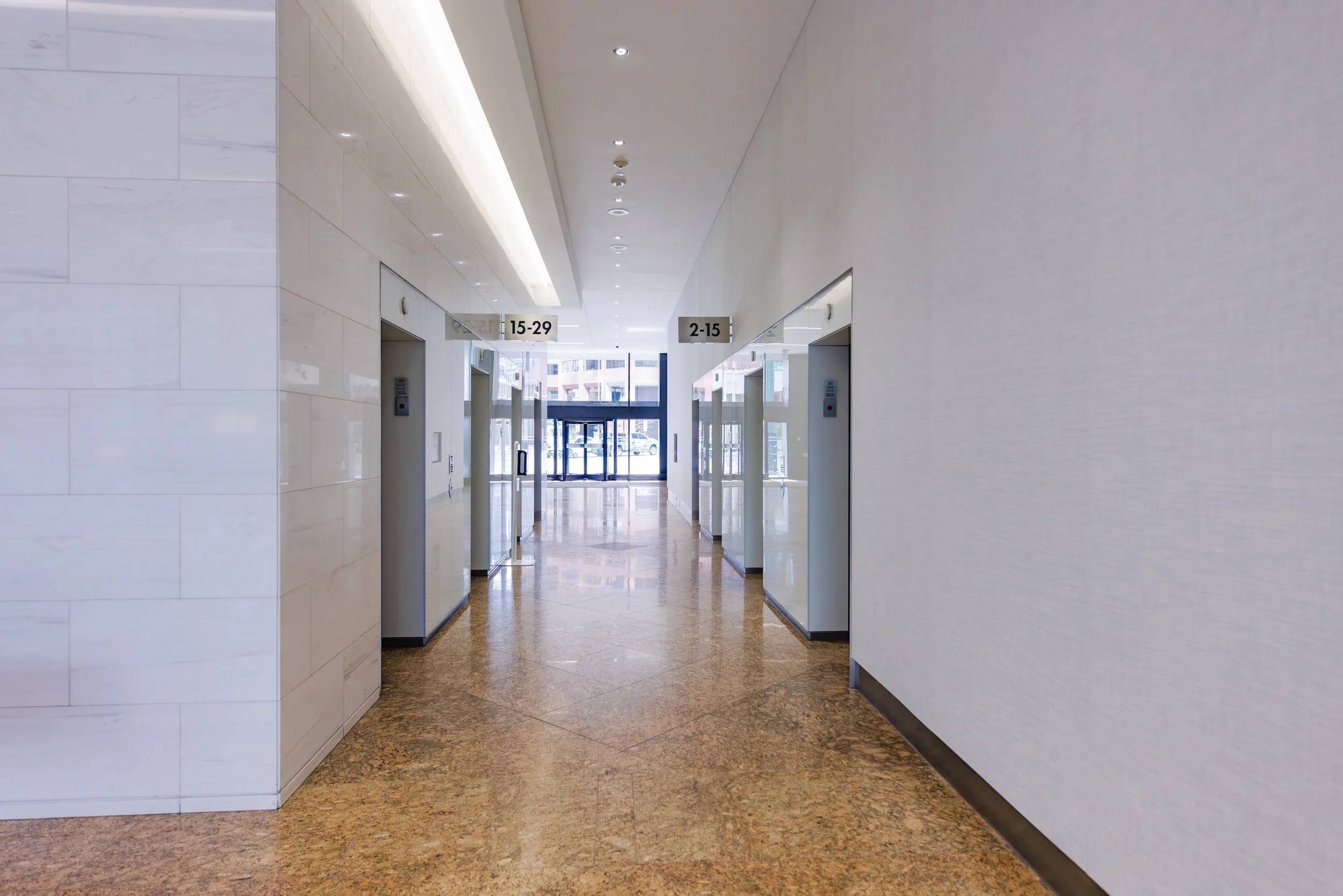 Corridor with two elevators on either side, digital floor indicators above each elevator, and glass doors at the end showing the outside street.