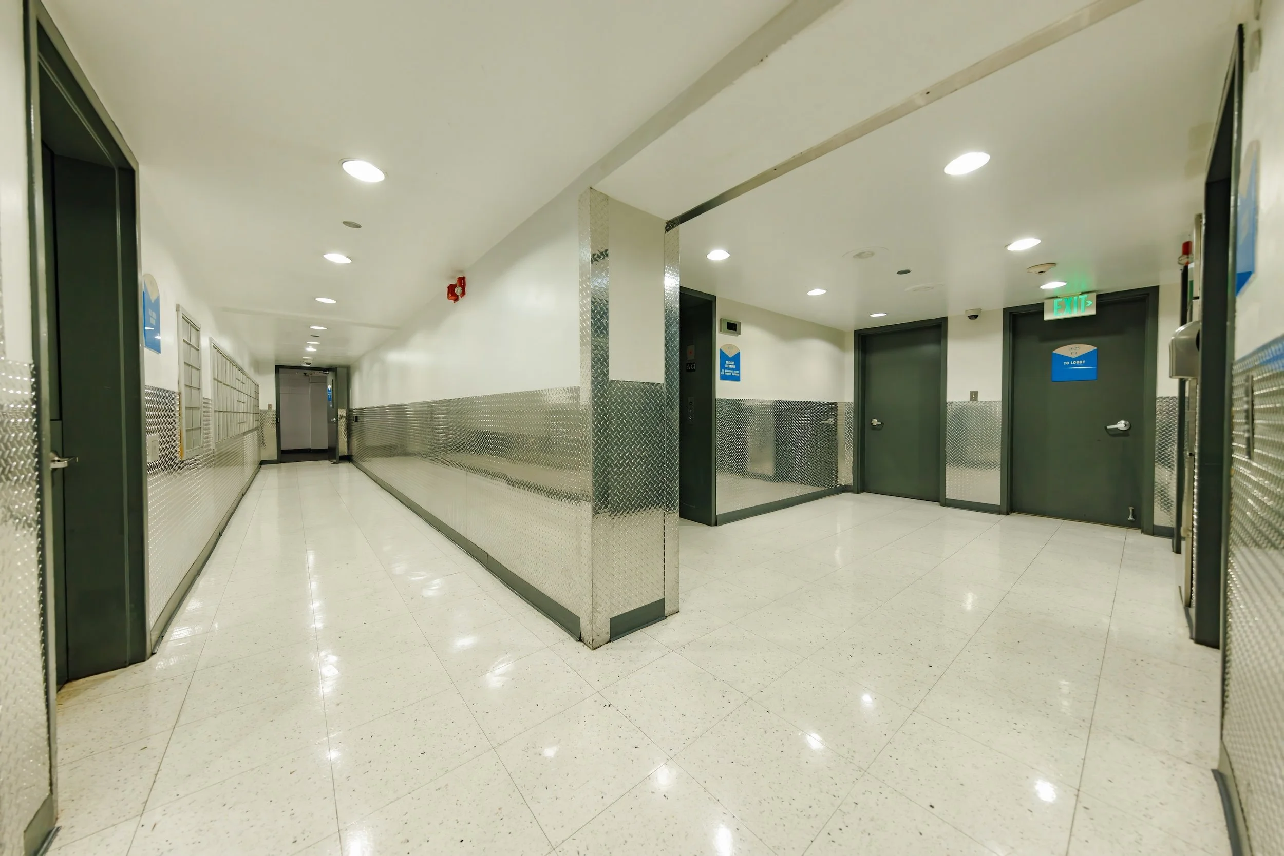 Empty hallway in a building with lockers on the left and doors on the right, illuminated by ceiling lights.