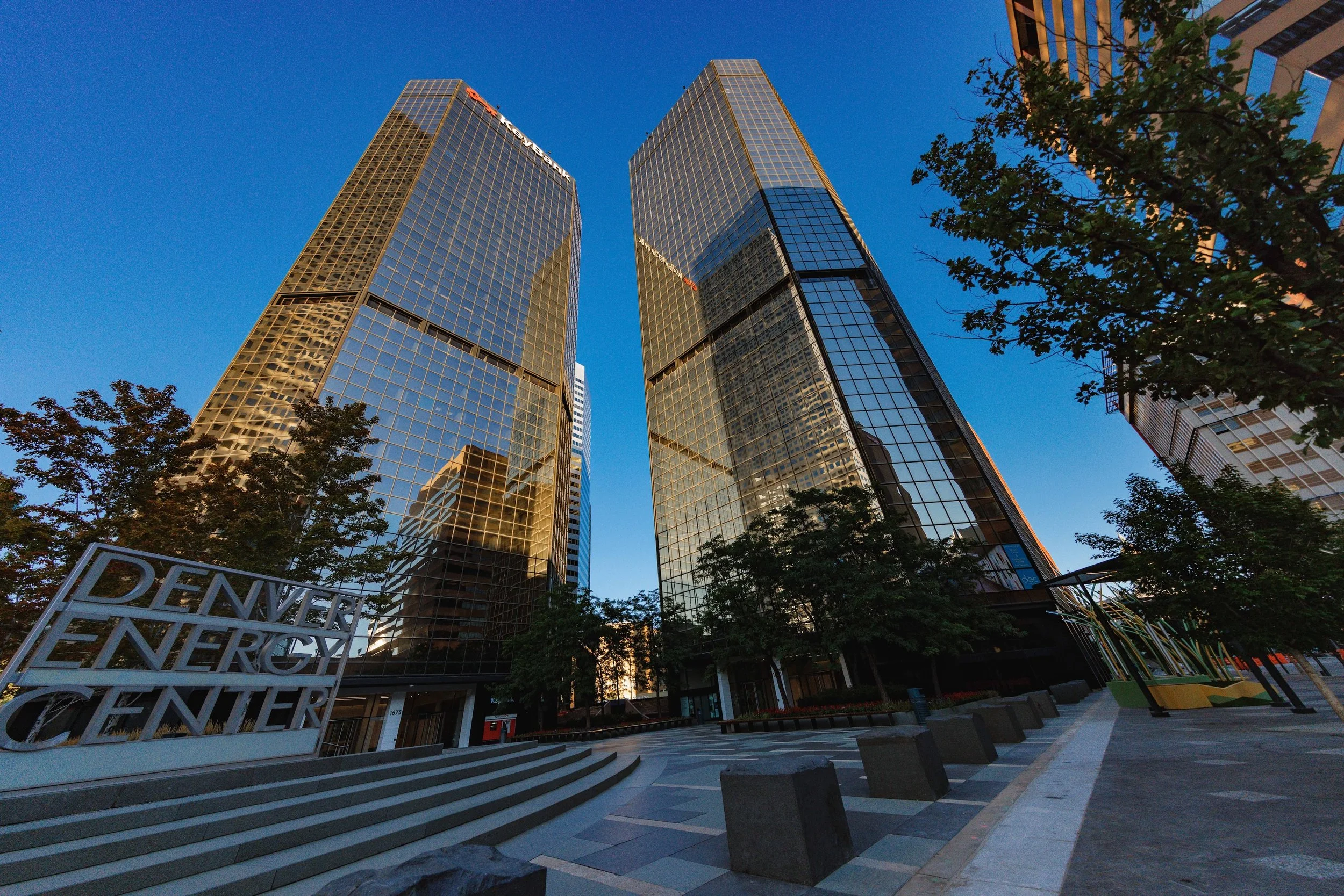 Tall glass skyscrapers reflecting the blue sky, with trees and steps in the foreground, and a sign that reads Denver Energy Center.