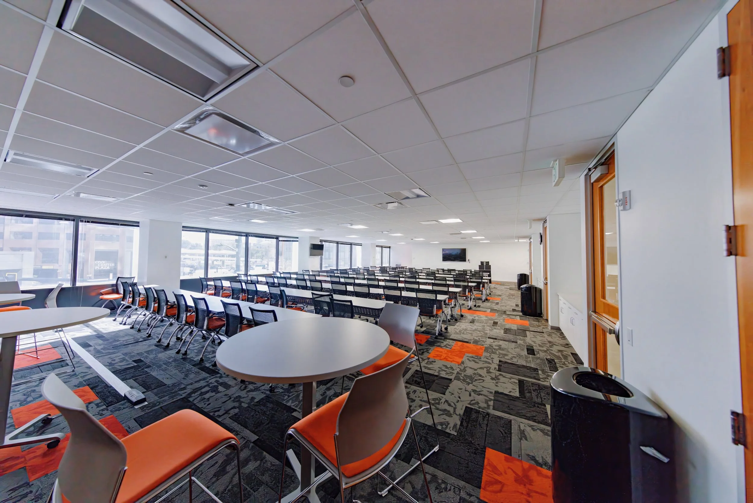 A spacious conference room with multiple rows of black office chairs and long tables, large windows providing natural light, and some round tables with orange-cushioned chairs in the foreground.