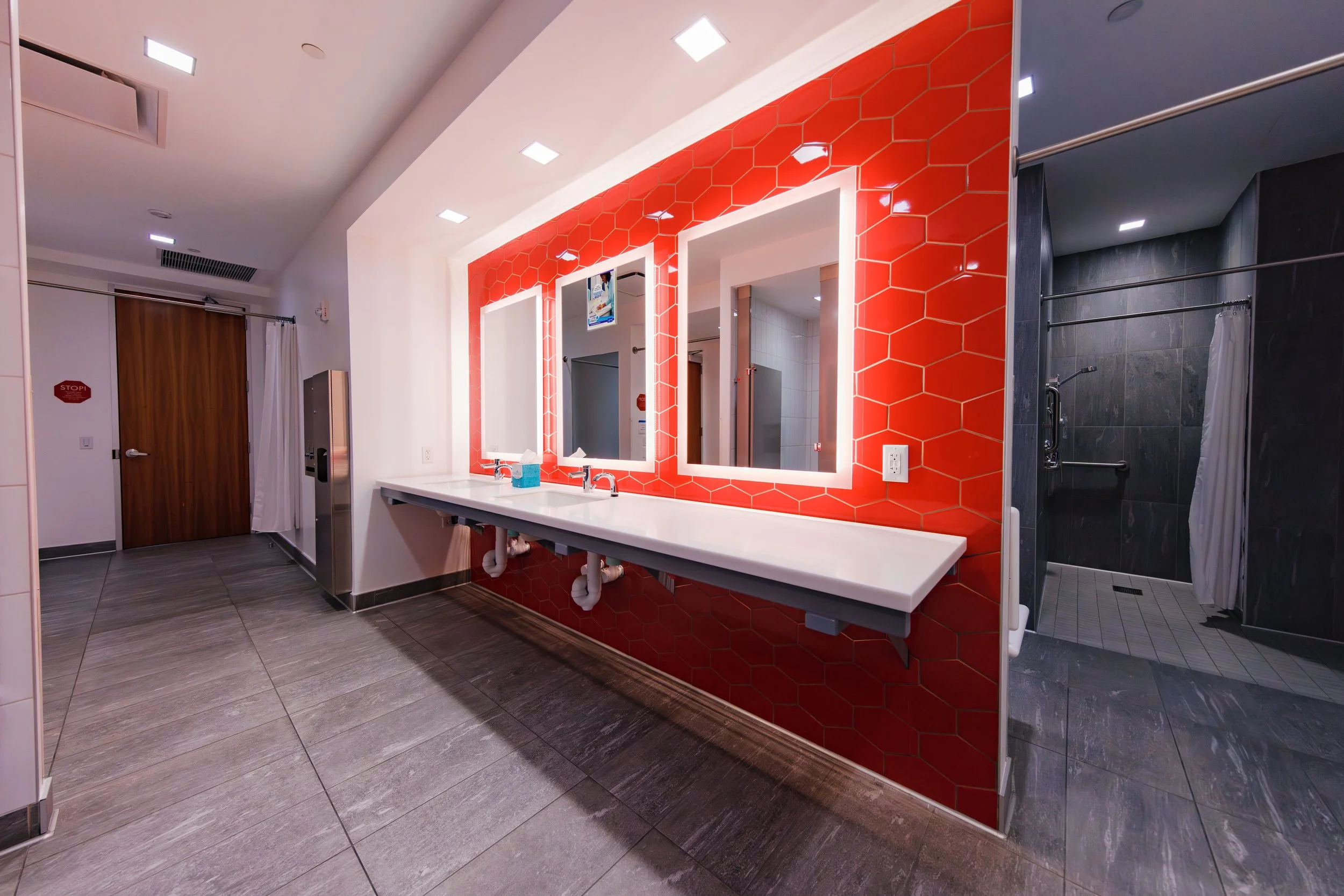 Public restroom with three backlit rectangular mirrors on a red hexagonal tiled wall, white countertop with sinks, and adjacent shower stall with dark wall tiles and curtain.