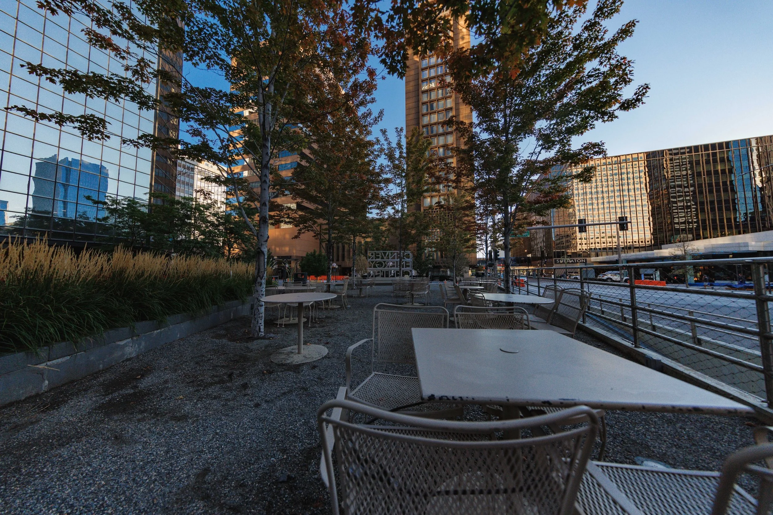 Empty outdoor patio with metal tables and chairs, trees, and modern glass buildings reflecting the sky and surroundings.
