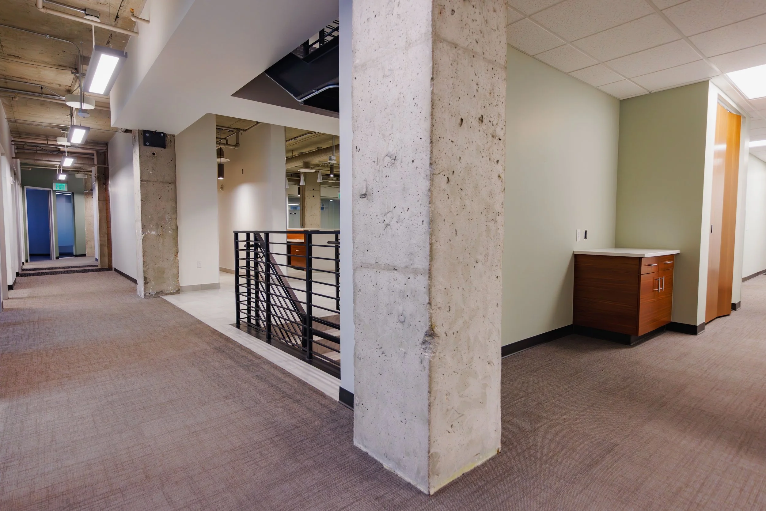 Interior view of an office hallway with a concrete pillar, carpeted flooring, and painted walls. There are ceiling lights, a staircase with a black railing, and several doorways and rooms visible in the background.