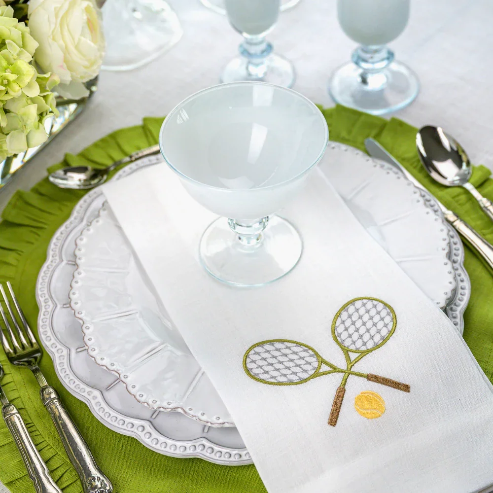 A formal dining table setting with a white plate, a white napkin with embroidered tennis rackets and a tennis ball, a clear glass goblet, and silverware on a green placemat, decorated with a floral centerpiece.