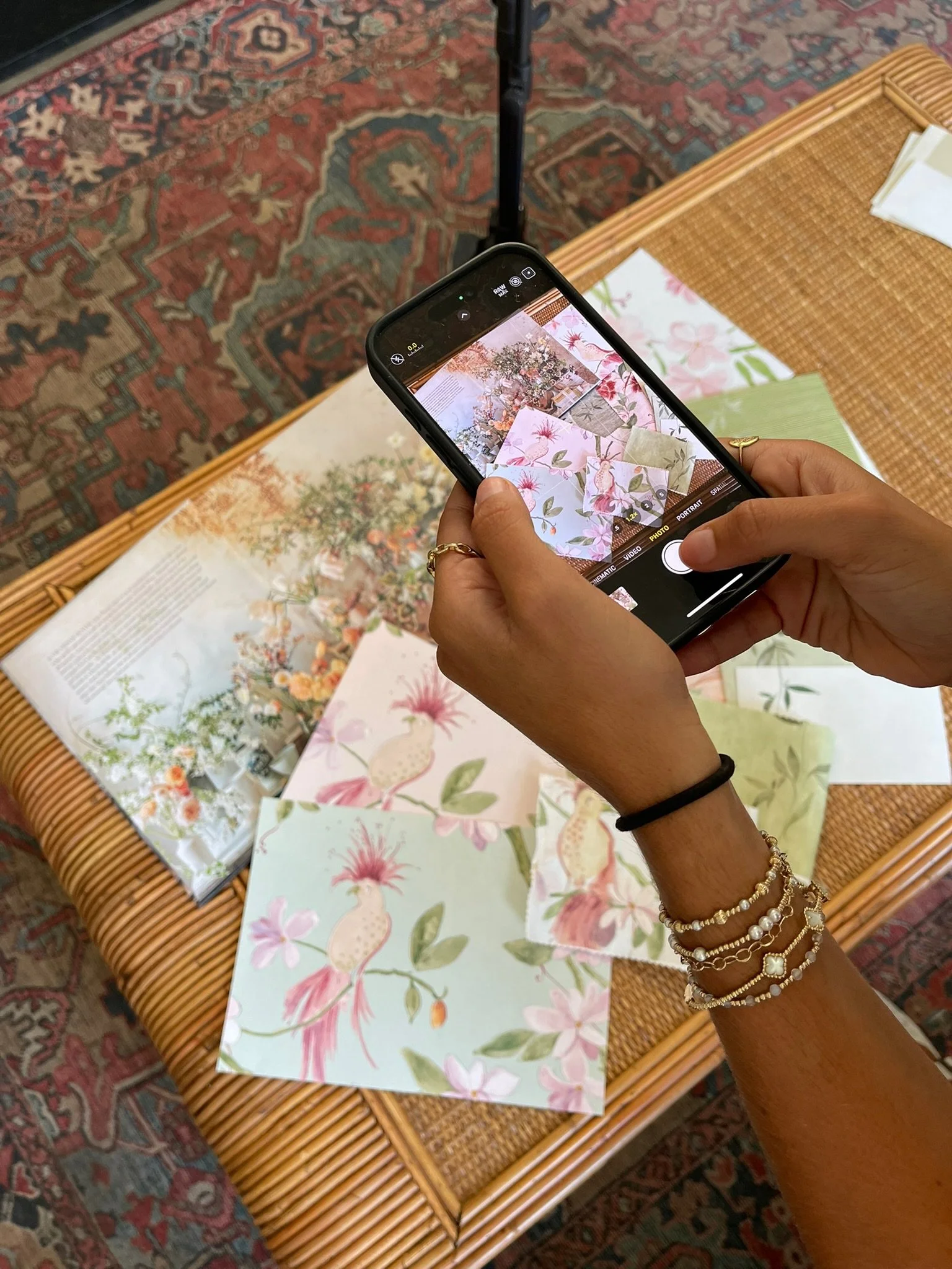 A person taking a photo of colorful floral and bird-themed papers on a wicker table with their smartphone. The papers feature illustrations of pink and white birds, flowers, and greenery.