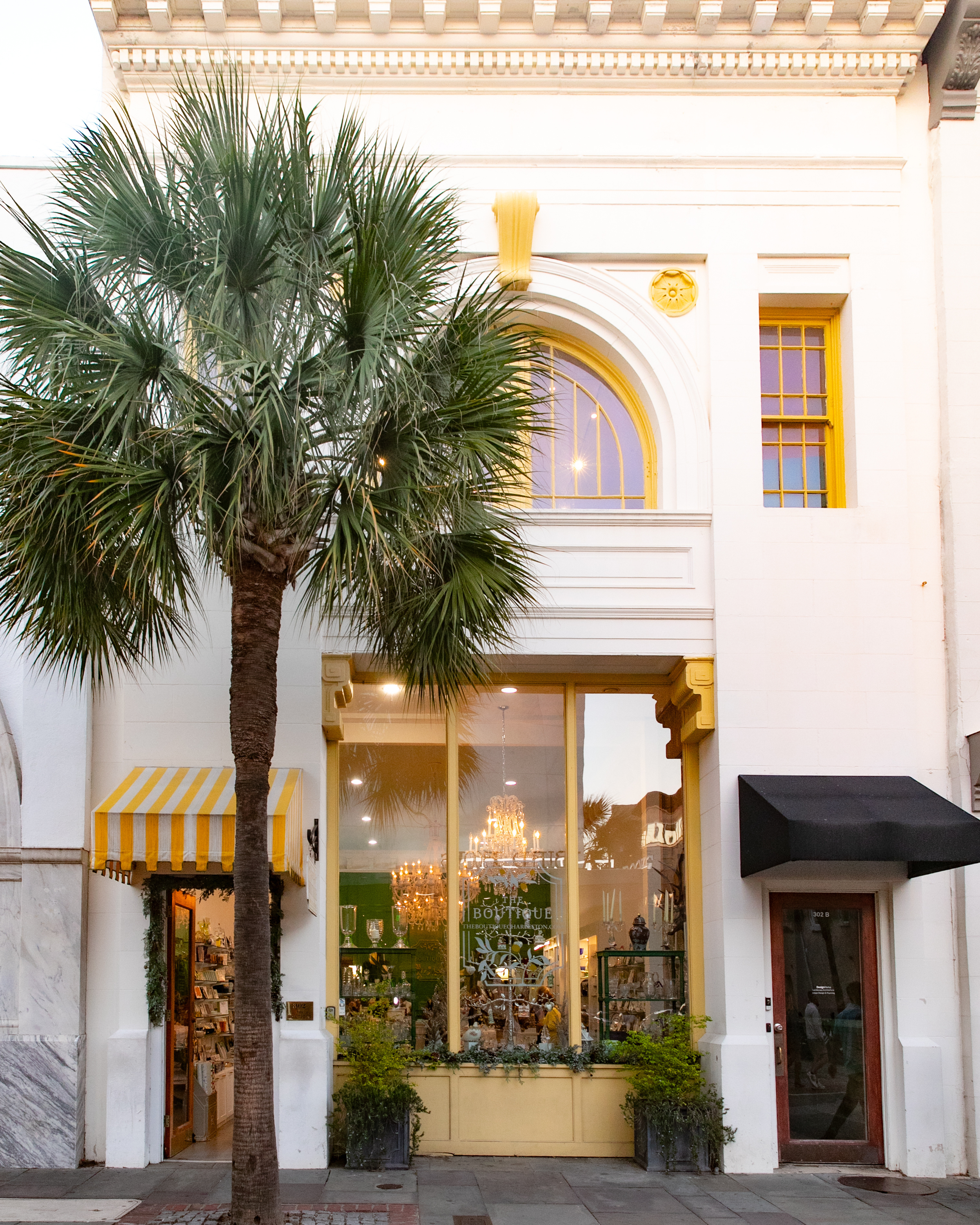 Front view of a boutique shop with a large window, yellow trim, and a black awning, featuring a tree in front.