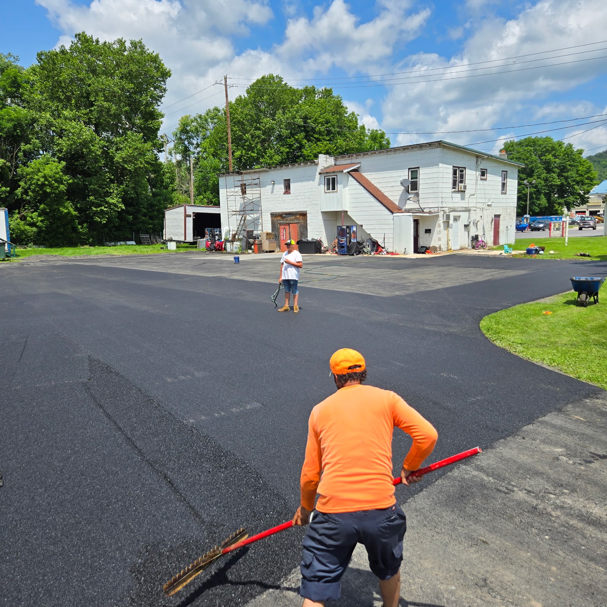 Two workers are resurfacing a parking lot with fresh black asphalt. One worker, wearing an orange shirt and cap, is raking the asphalt, while the other worker, wearing a white shirt and yellow cap, stands further back in the parking lot.
