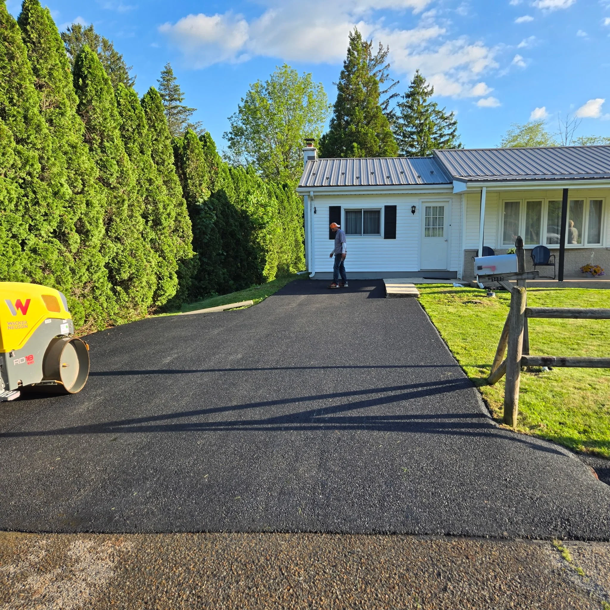 A newly paved black asphalt driveway leading to a white house, with a person standing on the driveway, a yellow asphalt roller on the left side, and a wooden fence on the right. The sky is blue with some clouds, surrounded by green trees.