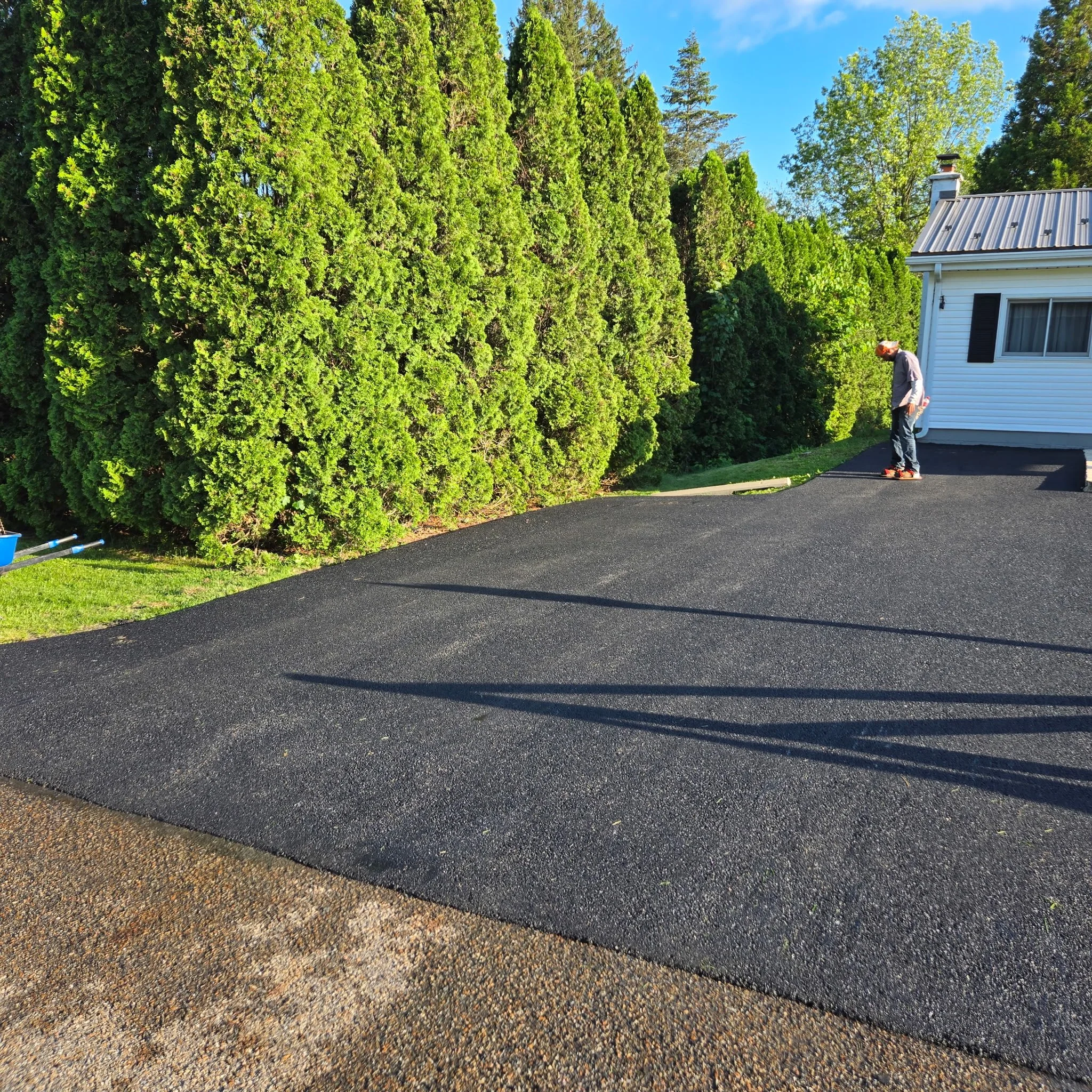 A person roller skating on freshly paved black asphalt driveway next to a white house, with tall green hedges and trees under a blue sky.