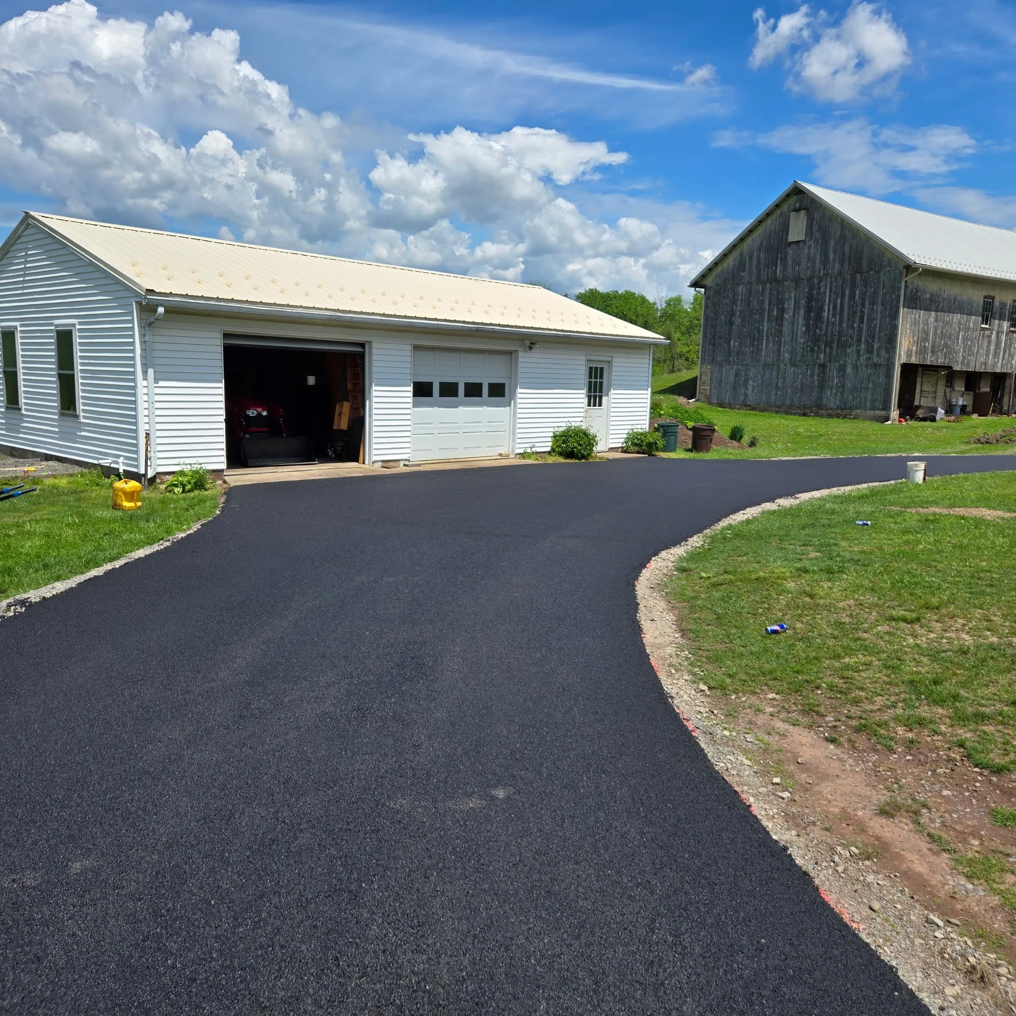 Newly paved black asphalt driveway curving past a white garage building with a open door, leading to a rural area with cloudy blue sky, green grass, and an older weathered barn in the background.