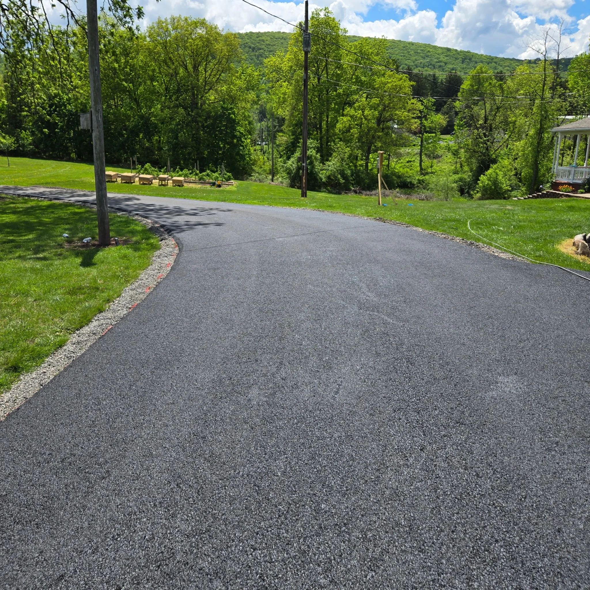 A freshly paved asphalt road in a rural area with green grass and trees on both sides, utility poles with power lines, a partly cloudy sky, and a hill in the background.