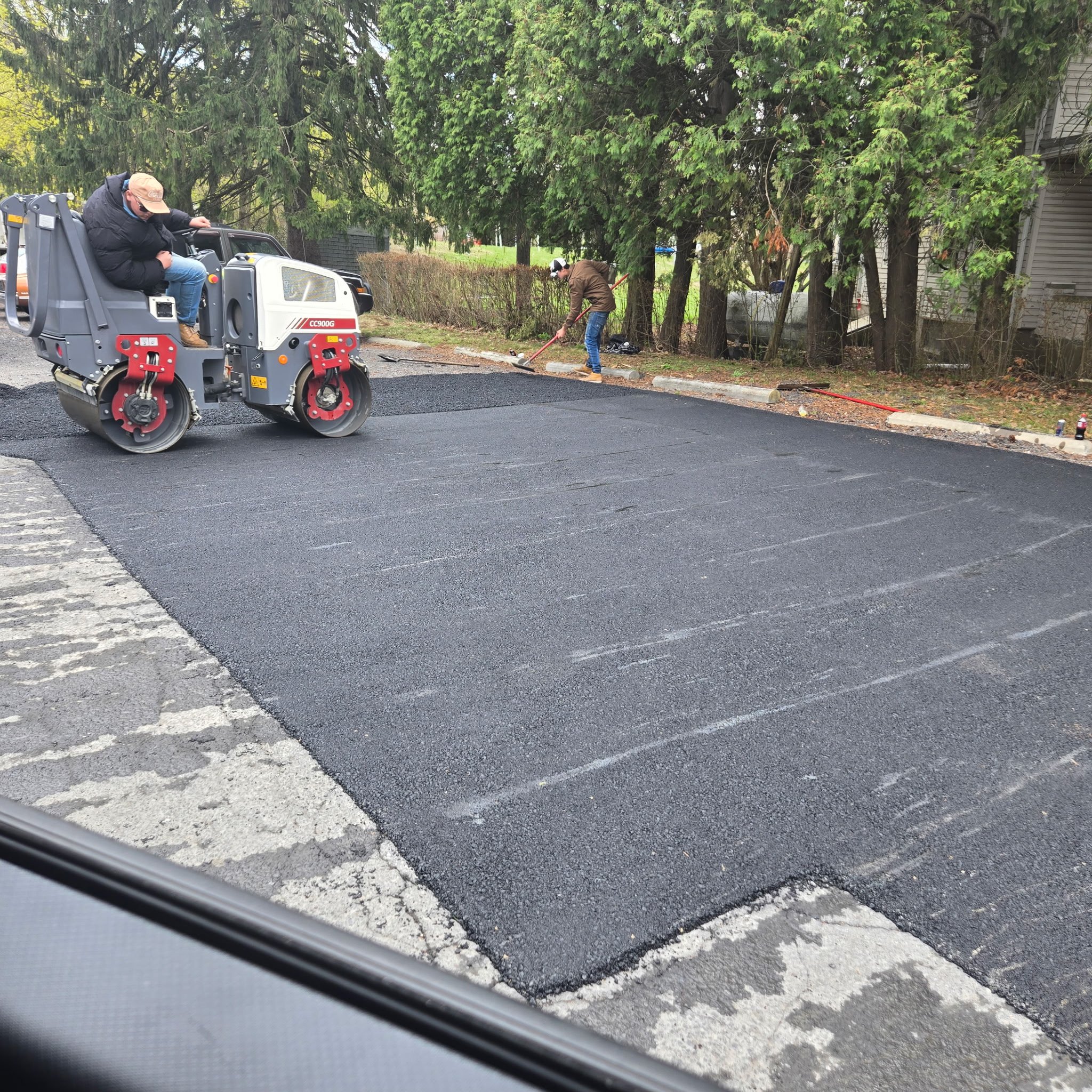 Workers are laying fresh asphalt on a road while a roller compactor smooths the surface. There are trees and a building in the background.