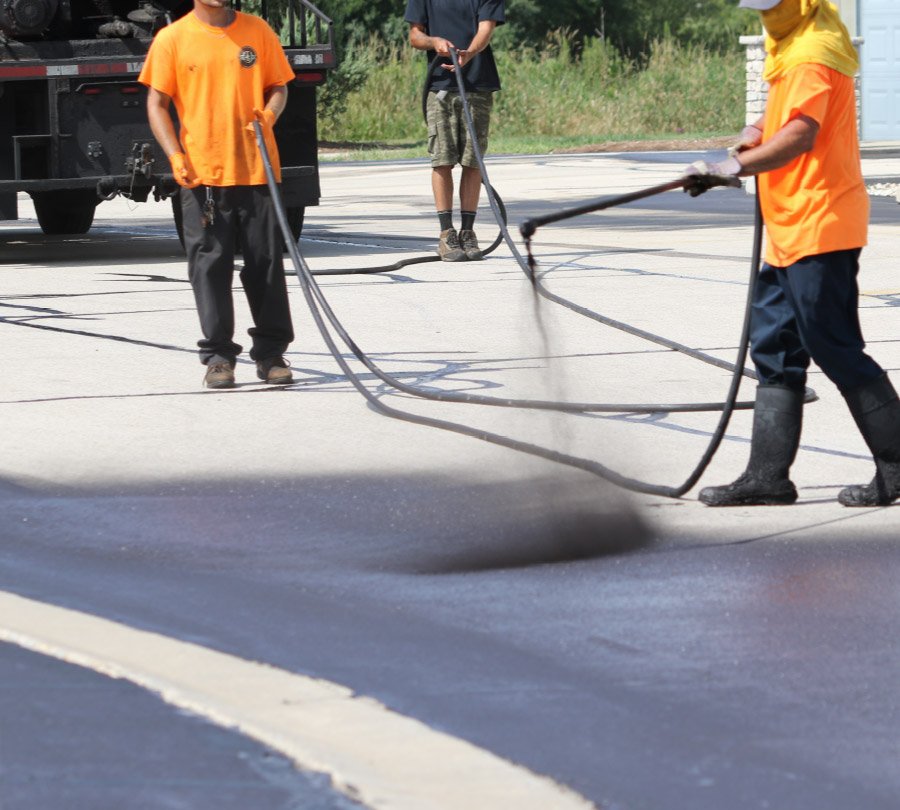 Workers are sealing a road with hot asphalt using a spray hose, wearing safety gear and orange shirts.