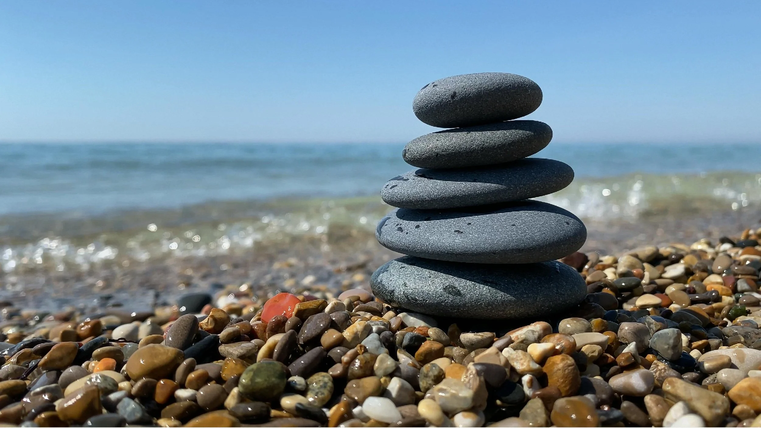 Stacked gray stones on a pebbled beach with the ocean and blue sky in the background.