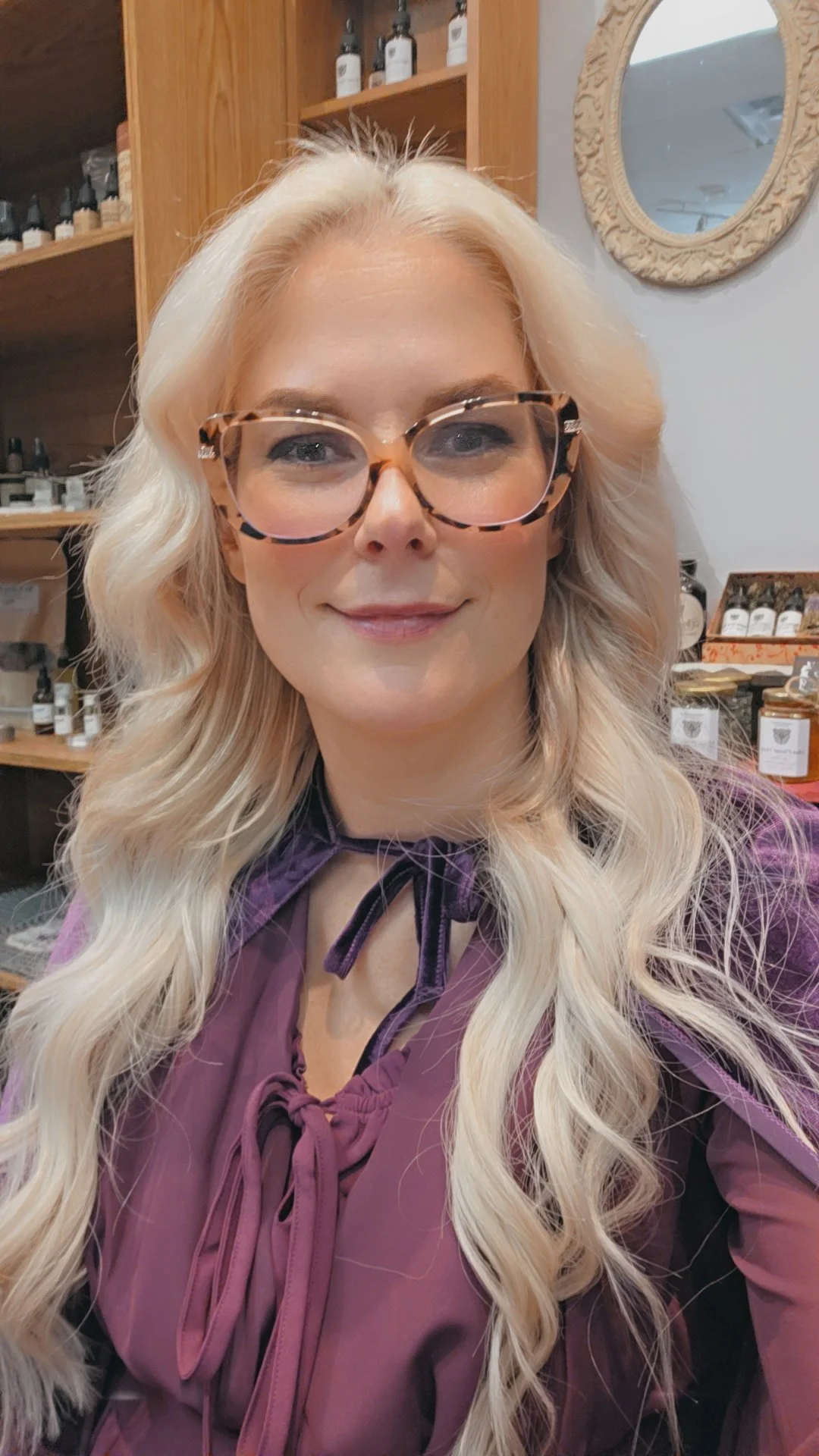A woman with long blonde hair, wearing tortoiseshell glasses and a purple blouse, smiling inside a store with wooden shelves and various products in the background.