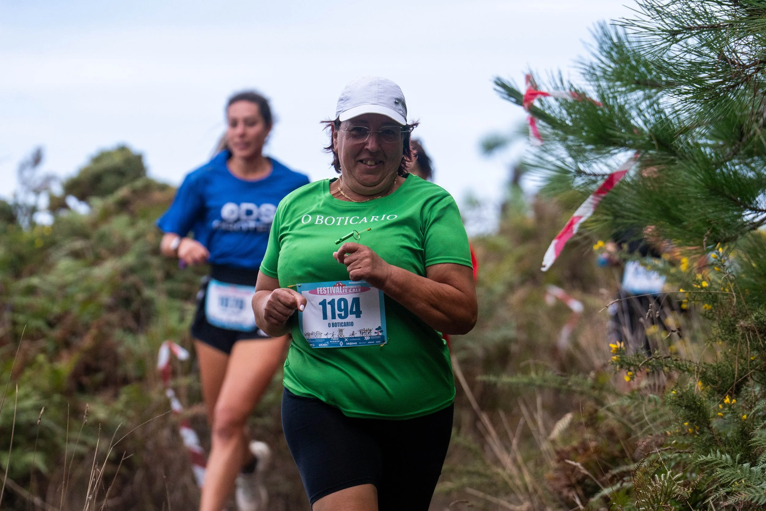 Two women running in a trail race through a natural outdoor setting with grass and trees, wearing athletic clothing and race bibs.