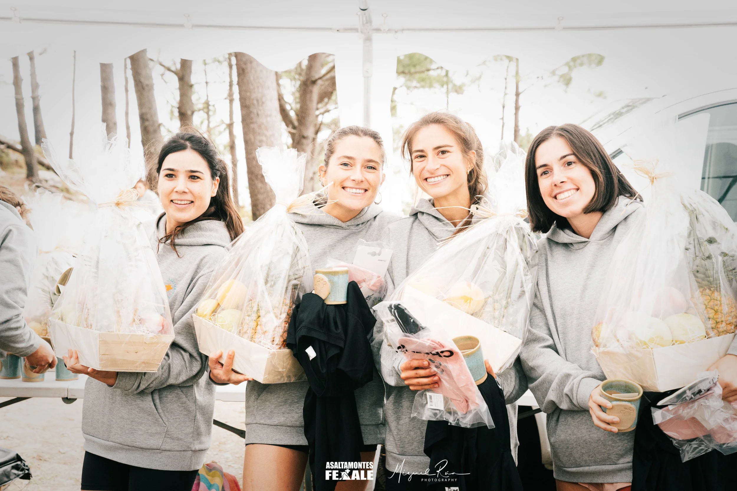 Four young women standing outdoors at a gathering, holding gift baskets with wrapped items and cups, smiling at the camera, wearing matching gray hoodies, with trees in the background.