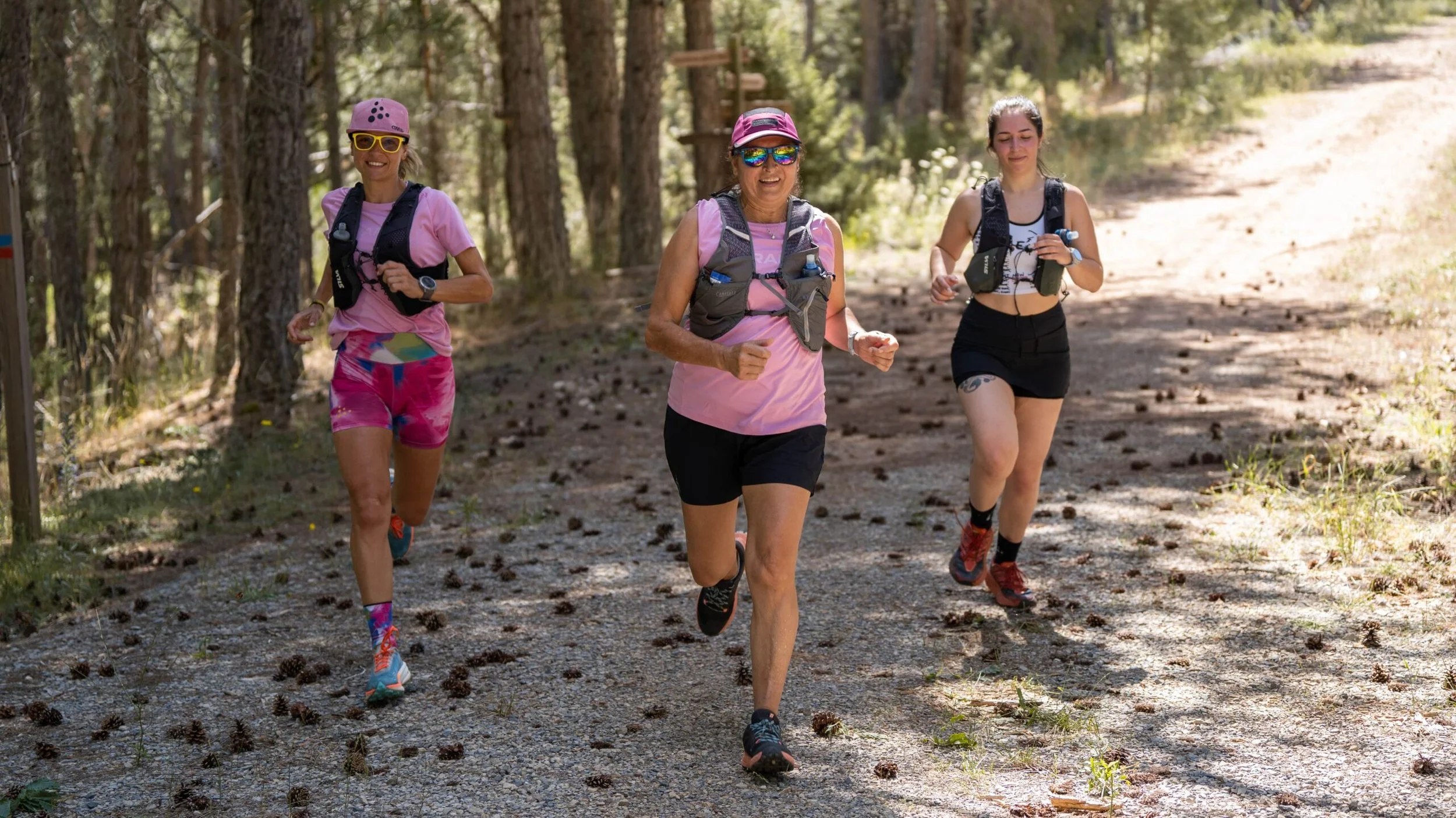 Three women trail running on a dirt path in a wooded area, wearing athletic gear and sunglasses.