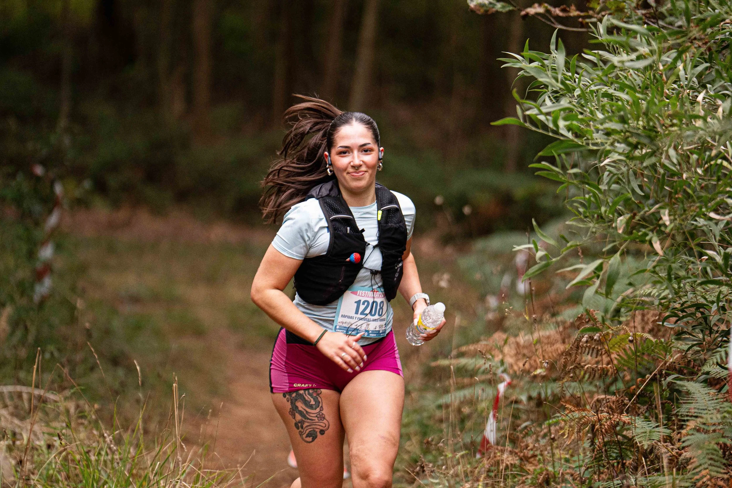 A woman running on a trail in a forest during a race, wearing athletic clothing, a backpack, and holding a water bottle.