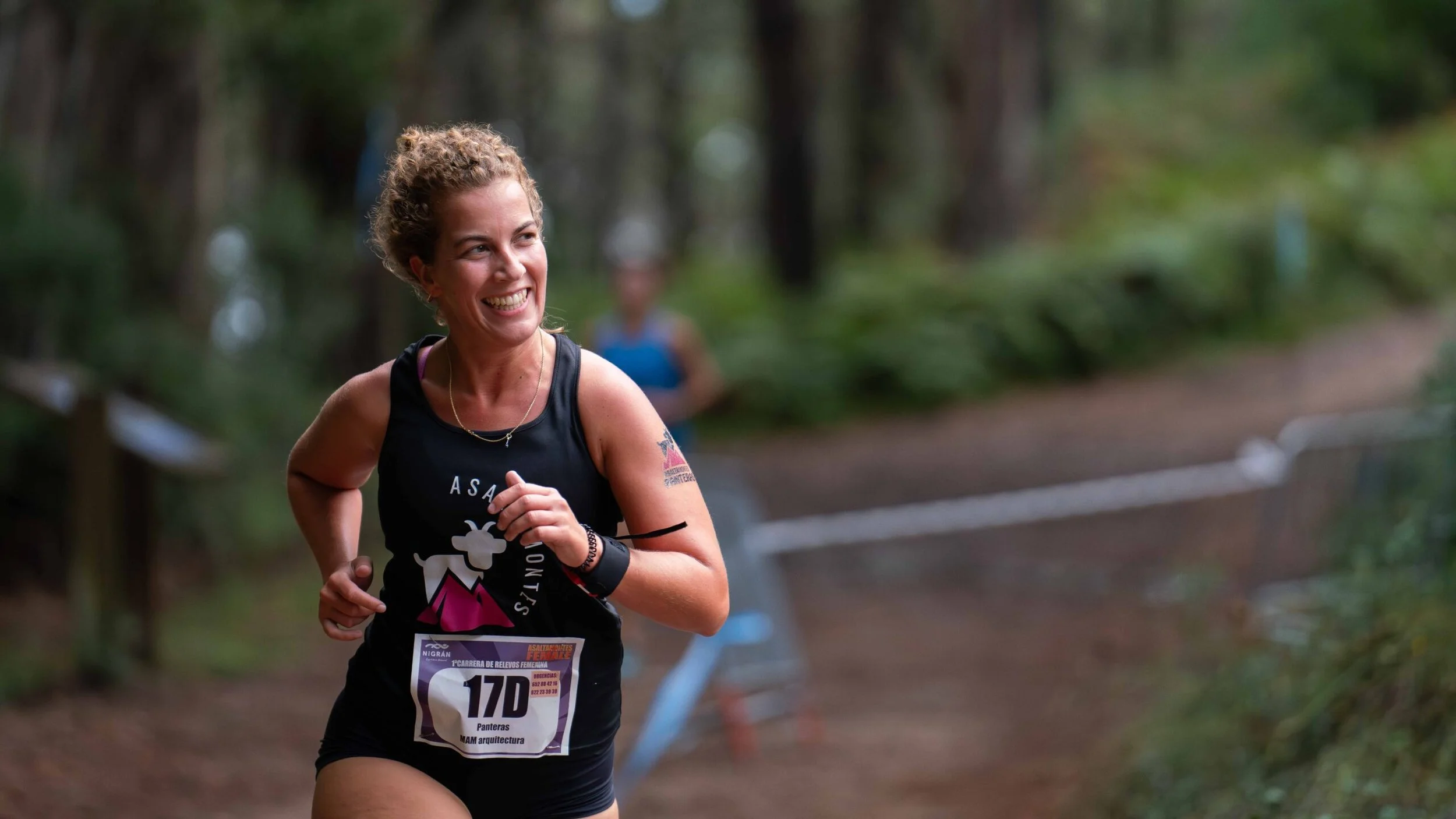 A smiling woman runs on a trail through a forest, wearing a black athletic tank top and shorts, with a race bib number 170.