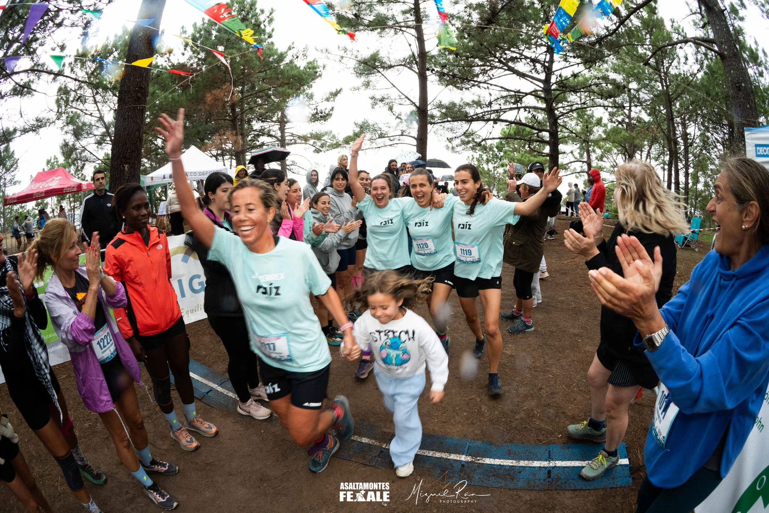 Group of women and children celebrating at a trail race or outdoor event in a wooded area, some wearing race bibs and matching teal shirts, with trees and banners in the background.