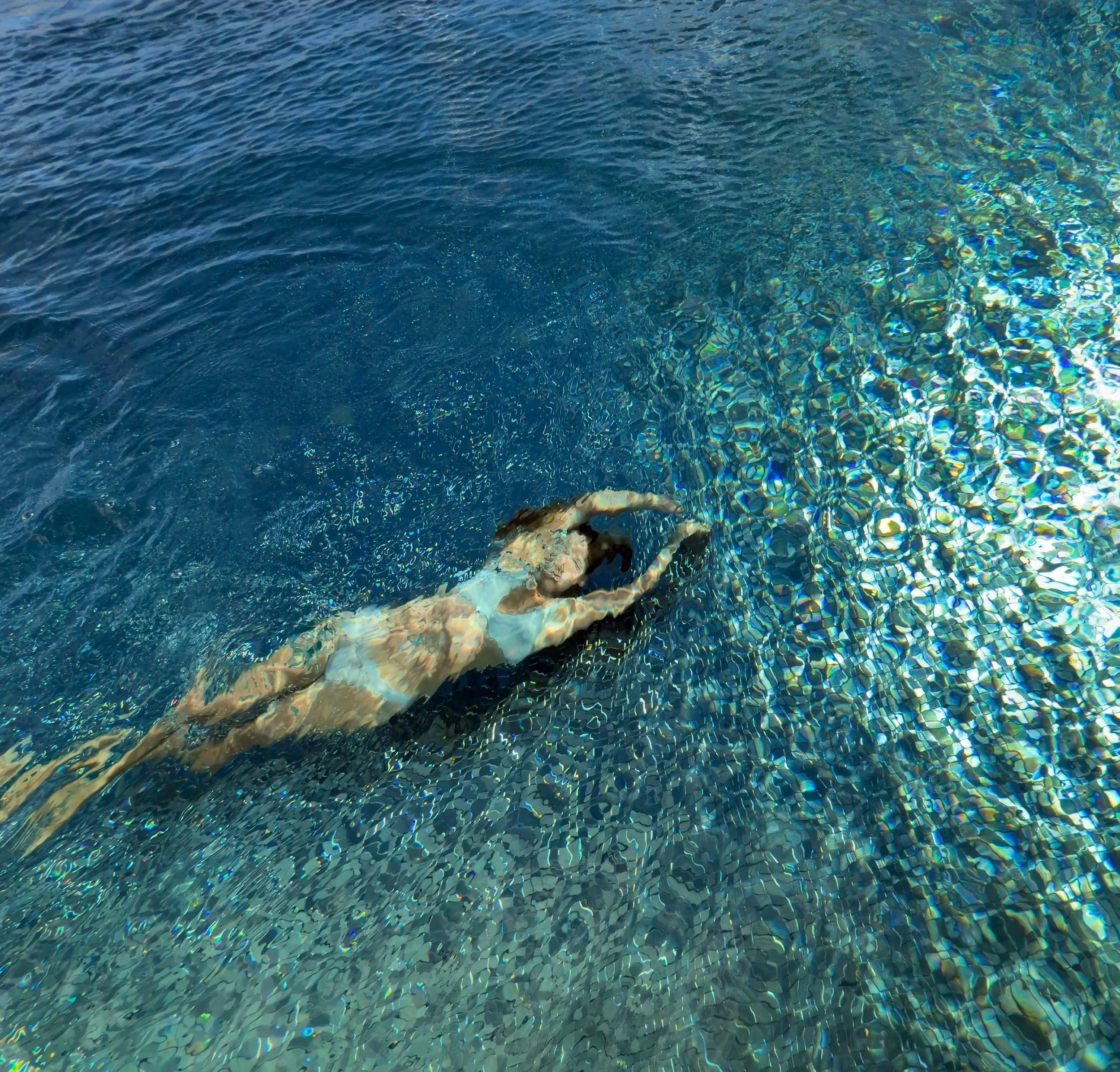 A person swimming underwater in a pool, making the 'OK' hand gesture while floating near the pool's shimmering tile floor.