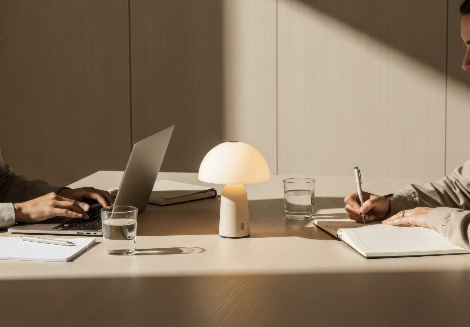 Two people sitting at a table with a lamp, two glasses of water, a closed laptop, notebooks, and pens, in a minimalist office setting.