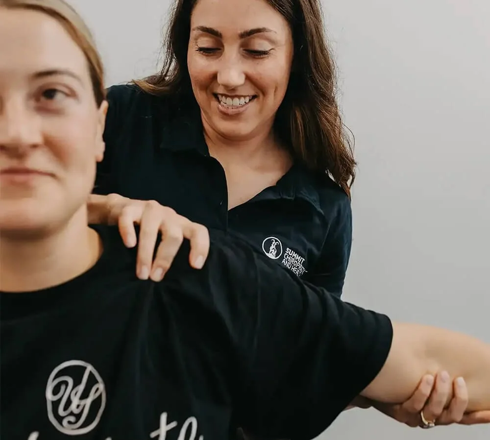 A woman providing physical therapy or chiropractic care to a young man, who is stretching his arm. Both are wearing black shirts with logos.