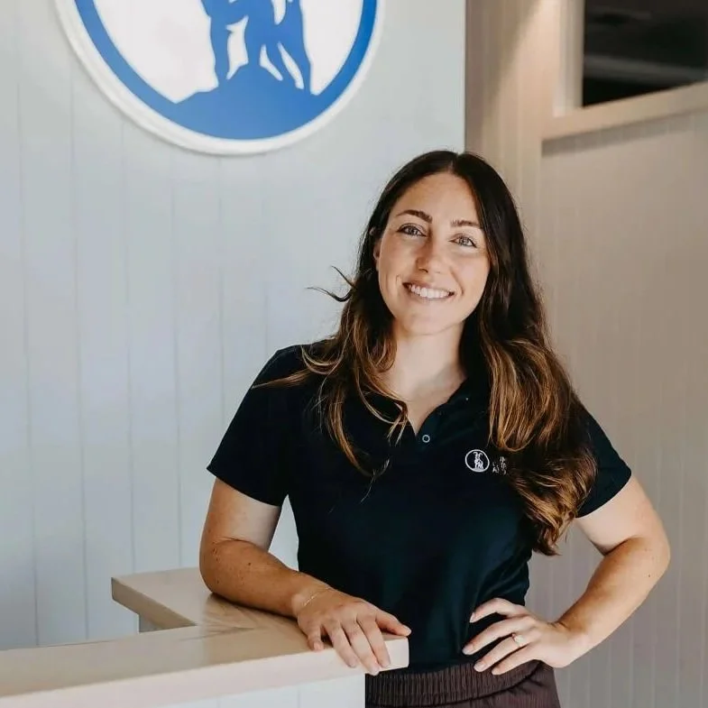 A smiling woman with long brown hair wearing a black polo shirt, standing behind a counter in front of a white wall with a blue logo.
