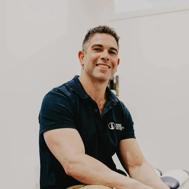 A smiling man with short dark hair, wearing a dark blue polo shirt, sitting in a room with a plain white wall.