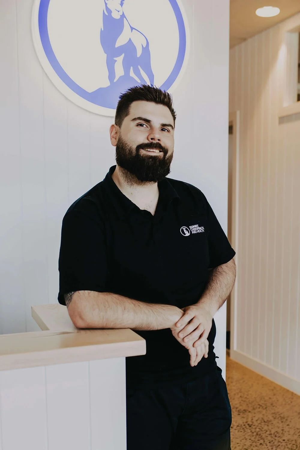 A man with a beard and dark hair, wearing a black polo shirt, standing indoors with a smile and crossed arms, in front of a wall with a circular logo featuring a figure in a dynamic pose.