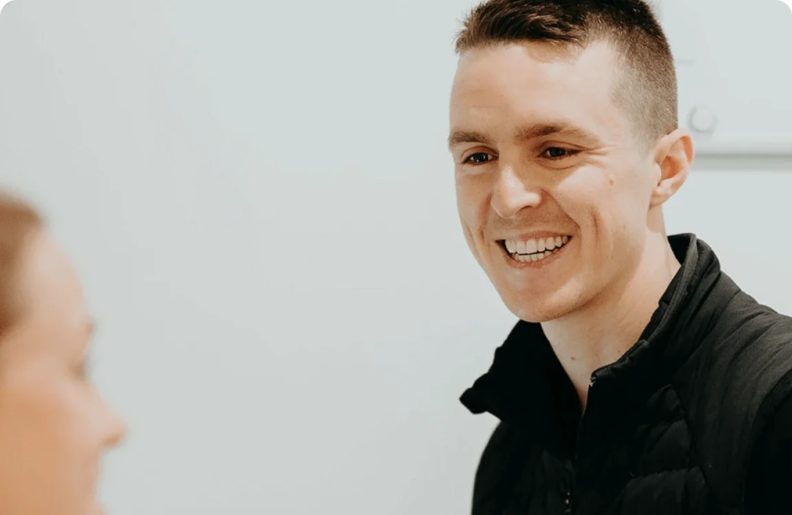 A young man with short brown hair smiling and talking to a woman with blonde hair in an indoor setting.