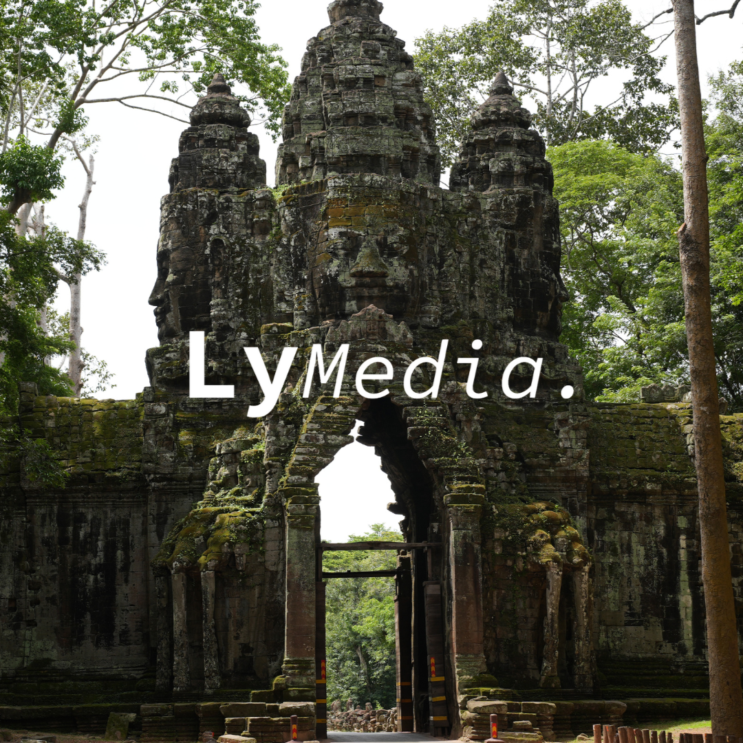 Ancient stone temple entrance with multiple tower-like structures with faces in Khmer style, surrounded by green trees in a jungle.