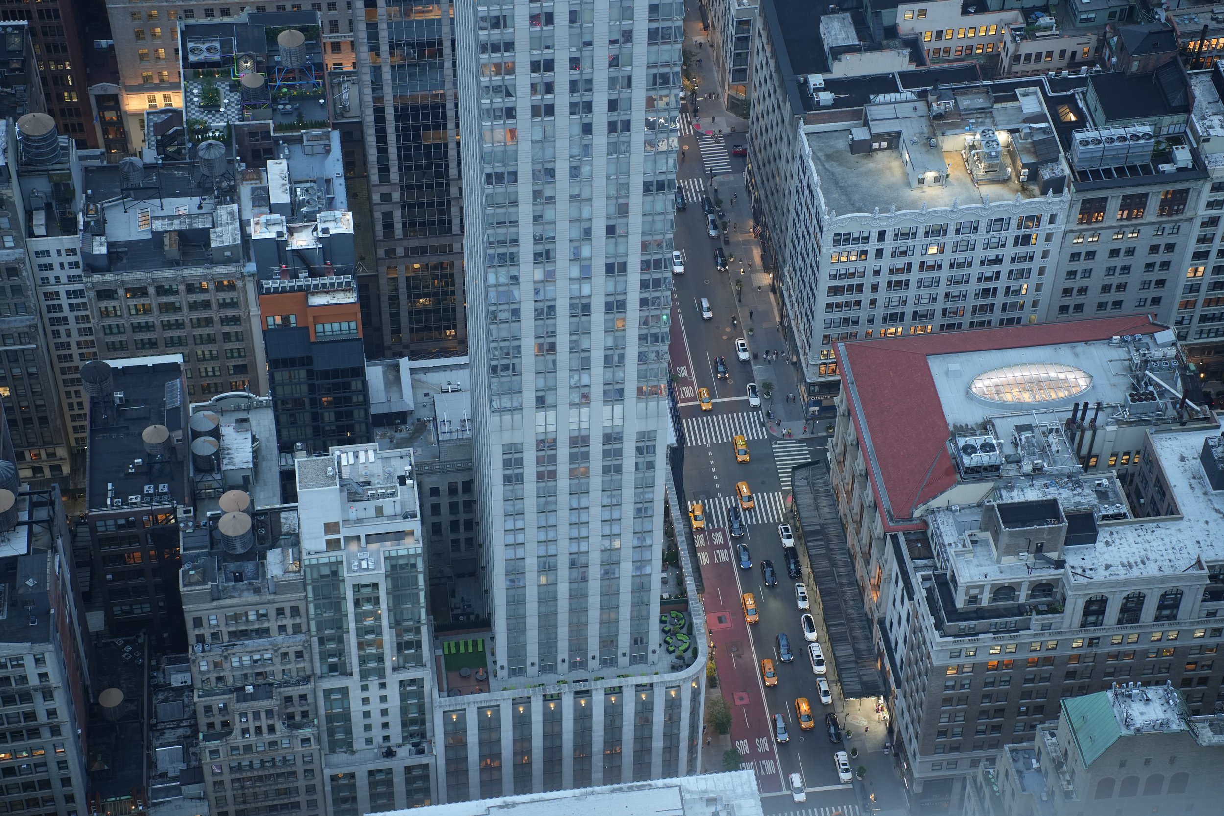 Aerial view of a city street surrounded by tall office buildings and skyscrapers with cars and taxis on the road, some with bus lanes marked 'Only Buses'.