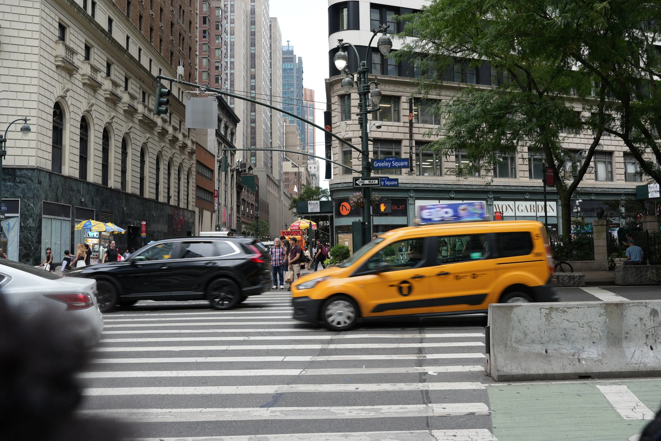 City street scene with yellow taxi, black SUV, and people crossing crosswalk in New York City. Buildings and street signs for Greeley Square visible.