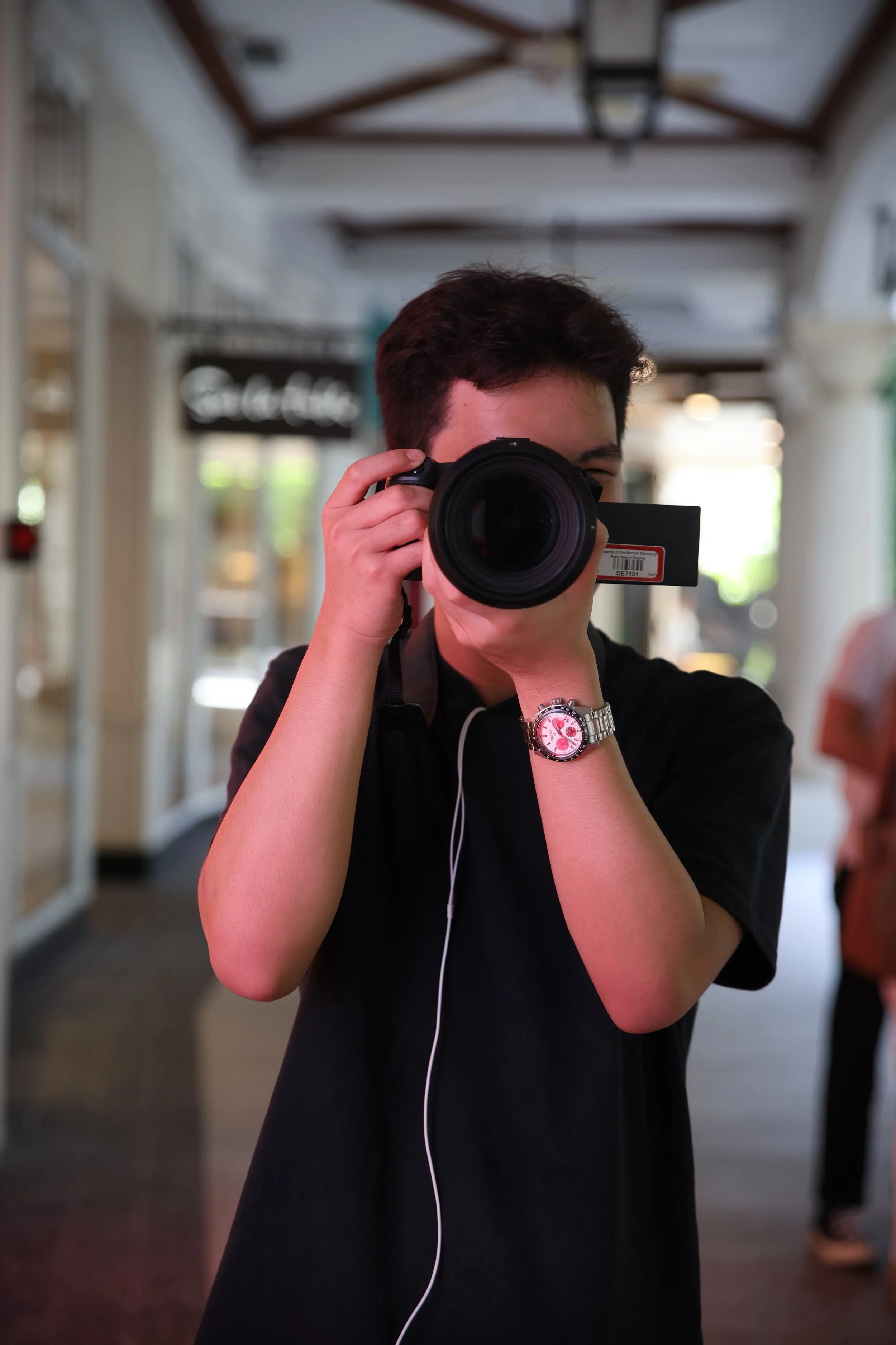 Person taking a photo with a camera in a shopping mall or indoor corridor, wearing a wristwatch, with a blurred background.