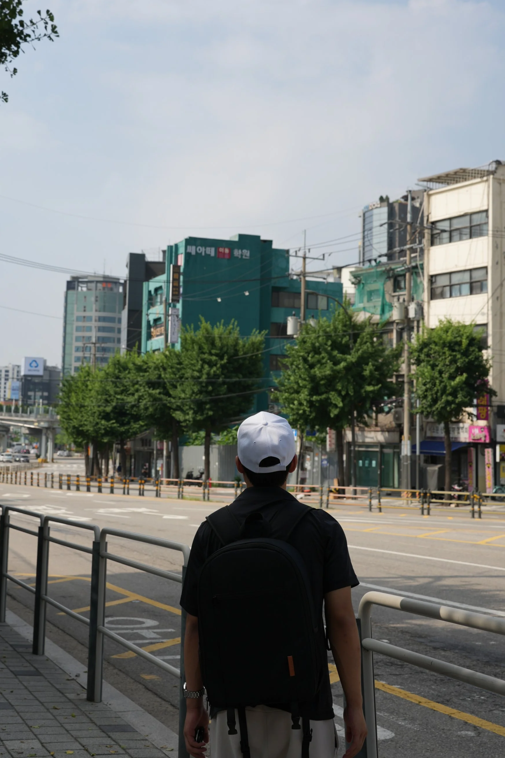 Person wearing a white cap, black shirt, and backpack standing at a city bus stop, looking across an empty street lined with trees and high-rise buildings.