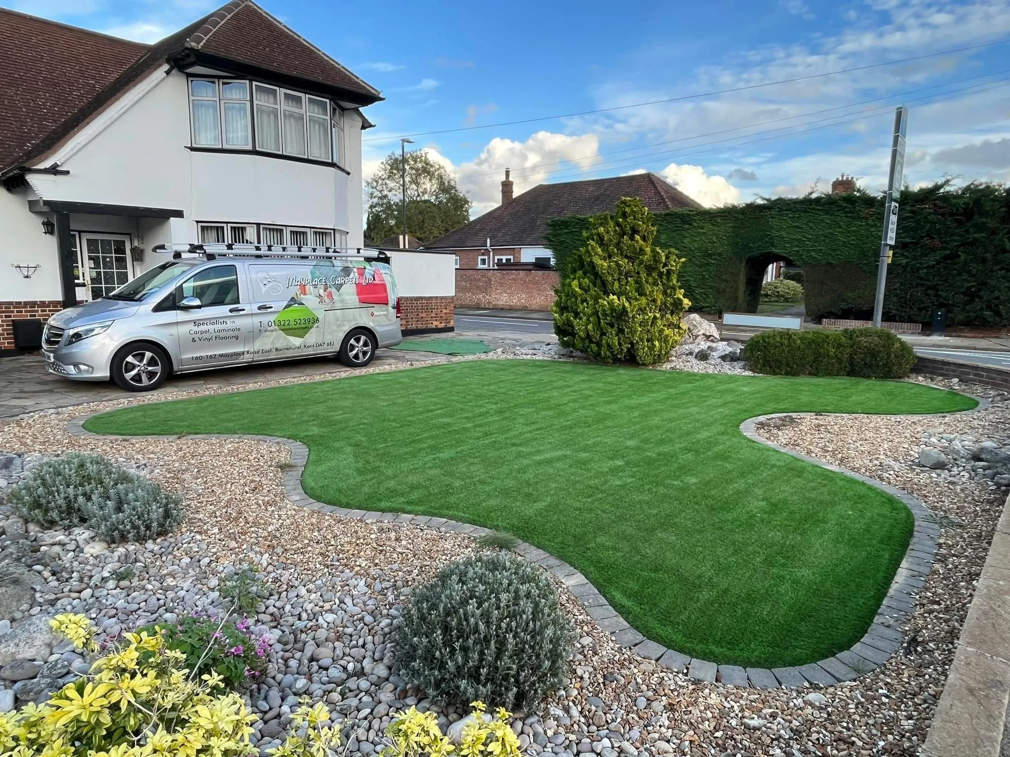 A small landscaped yard with a green artificial grass patch, surrounded by gravel and decorative plants, with a house and car parked nearby on a suburban street.