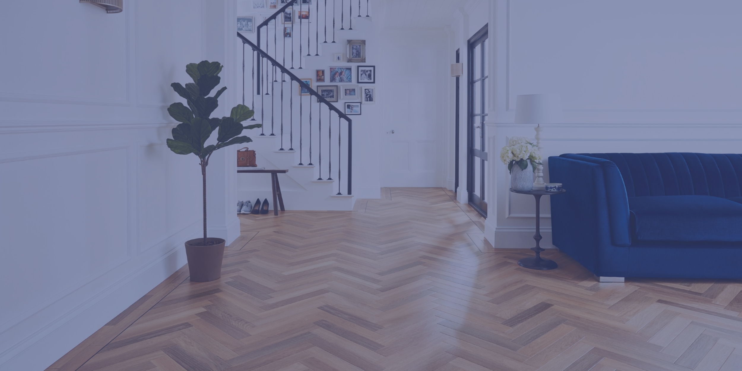 Interior of a modern living room with light-colored walls, wooden herringbone floor, large blue velvet sofa, side table with flowers, gallery wall near staircase, and entry door.