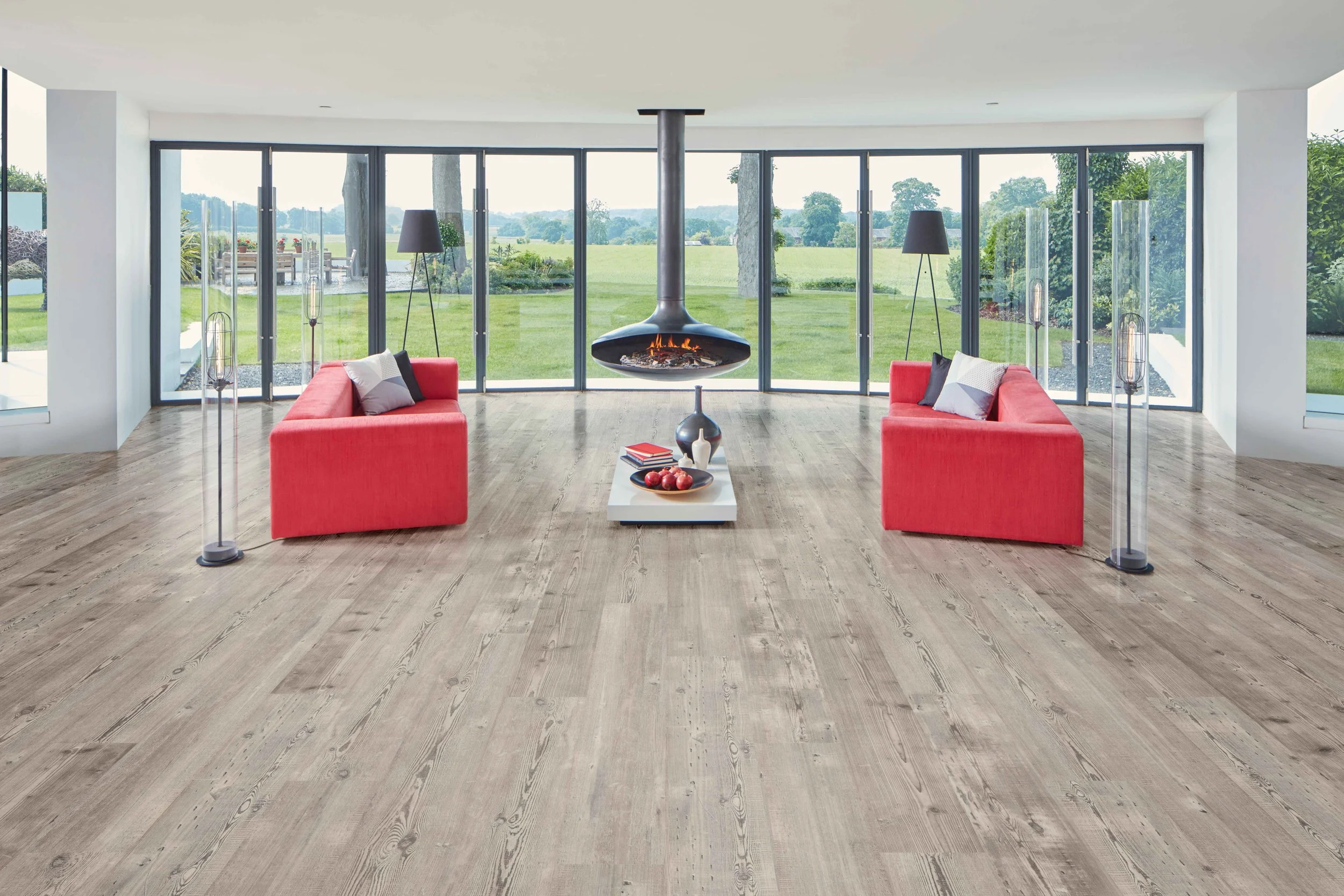 Modern living room with large floor-to-ceiling windows, two red sofas, a fireplace hanging from the ceiling, and a view of a green lawn outside.