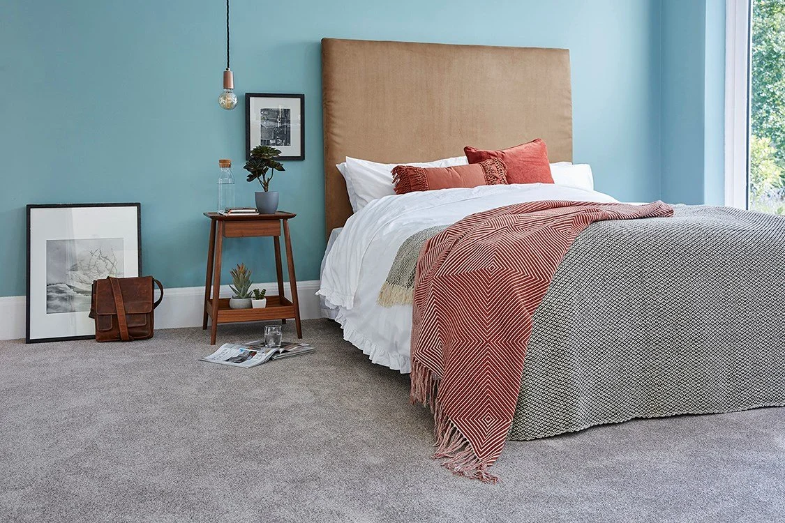 A neatly made bed with a tan upholstered headboard, white sheets, and red and beige pillows, beside a wooden nightstand with a potted plant and a water bottle, in a bedroom with blue walls, a large window, and minimal decor.