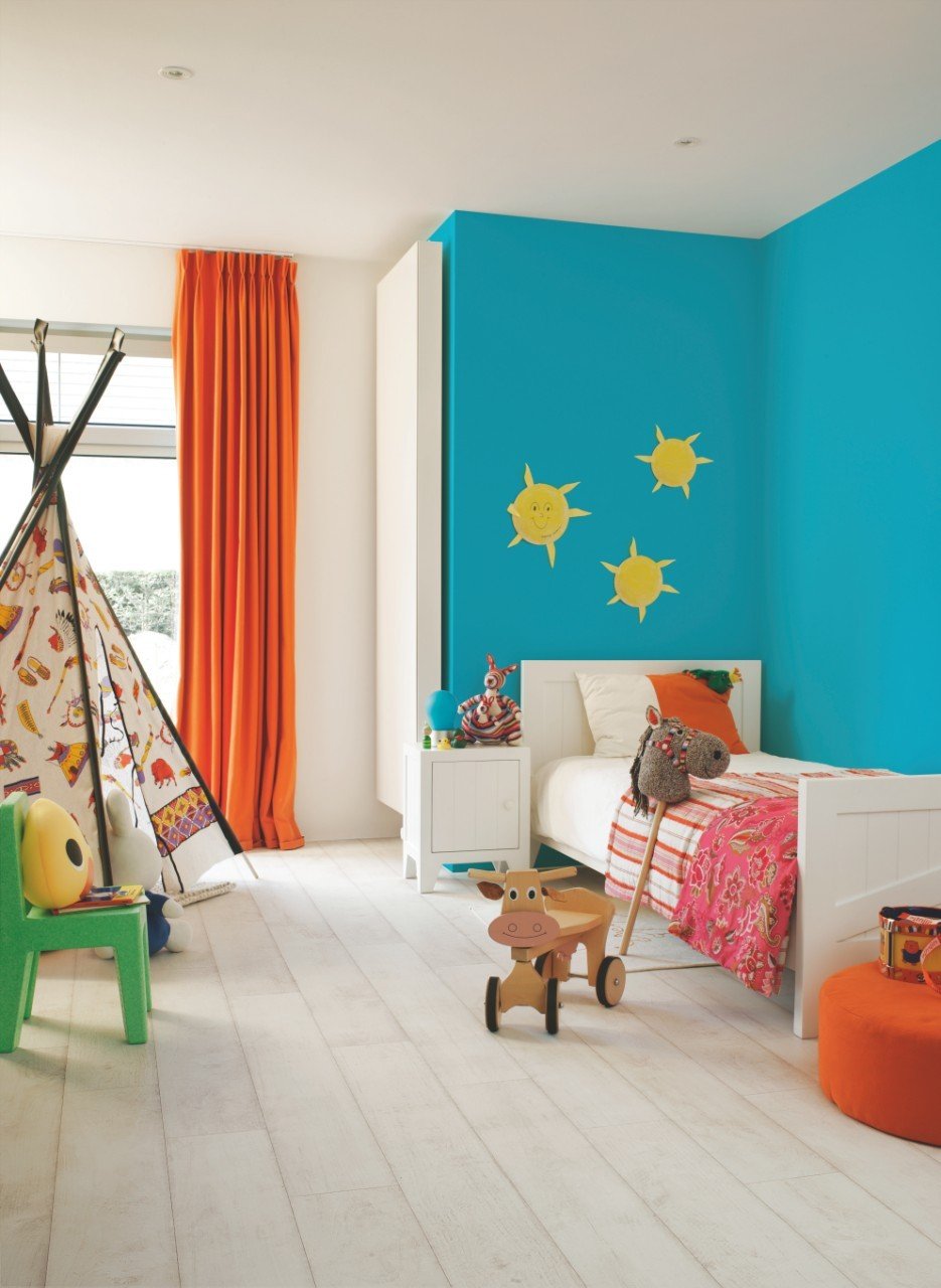 Colorful children's bedroom with a blue accent wall, decorated with painted suns, a white bed with colorful bedding and toys, an orange curtain, a teepee tent, and various toys on the floor.