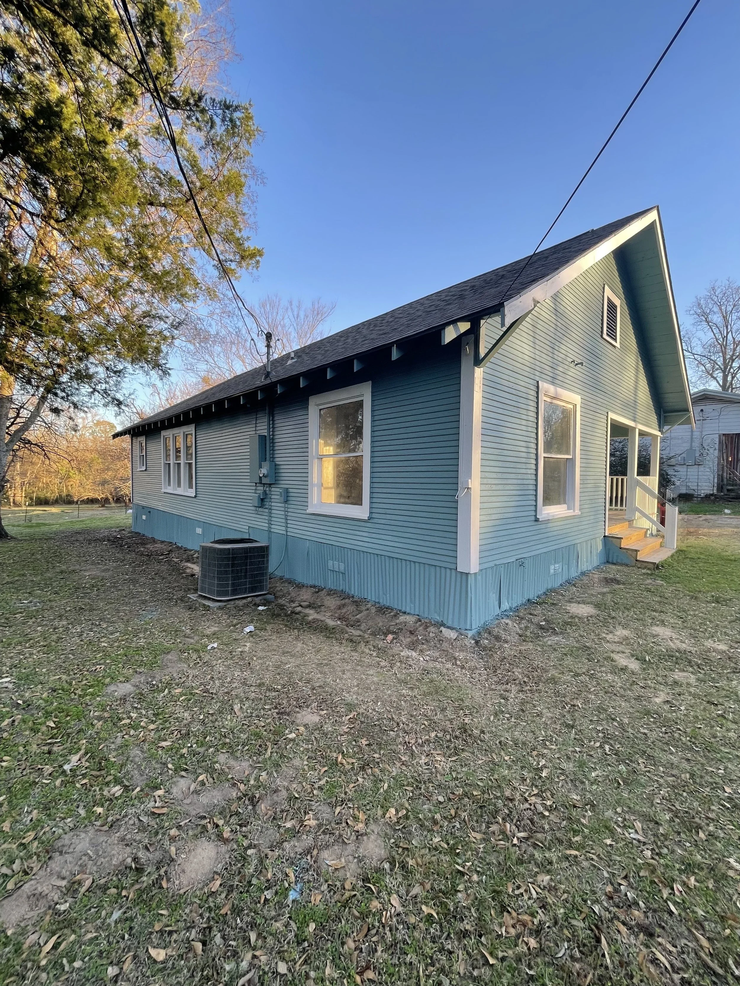 Blue single-story house with white trim, front steps, and small porch, surrounded by bare yard with some fallen leaves, under a clear blue sky.
