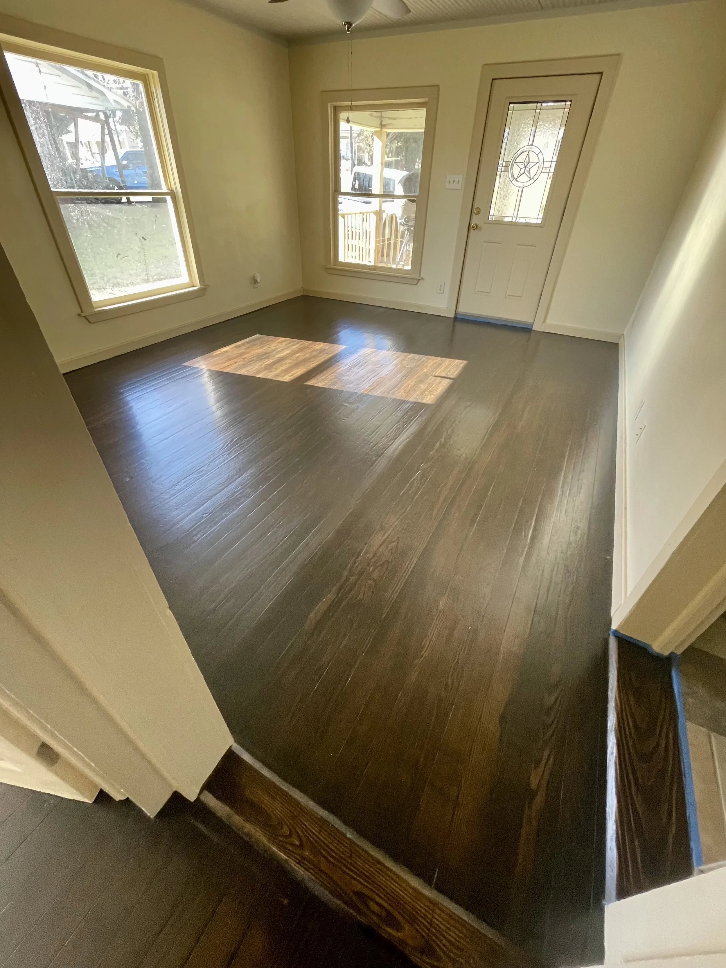 Empty living room with dark hardwood floors, cream walls, two large windows, and a front door with a decorative glass panel.
