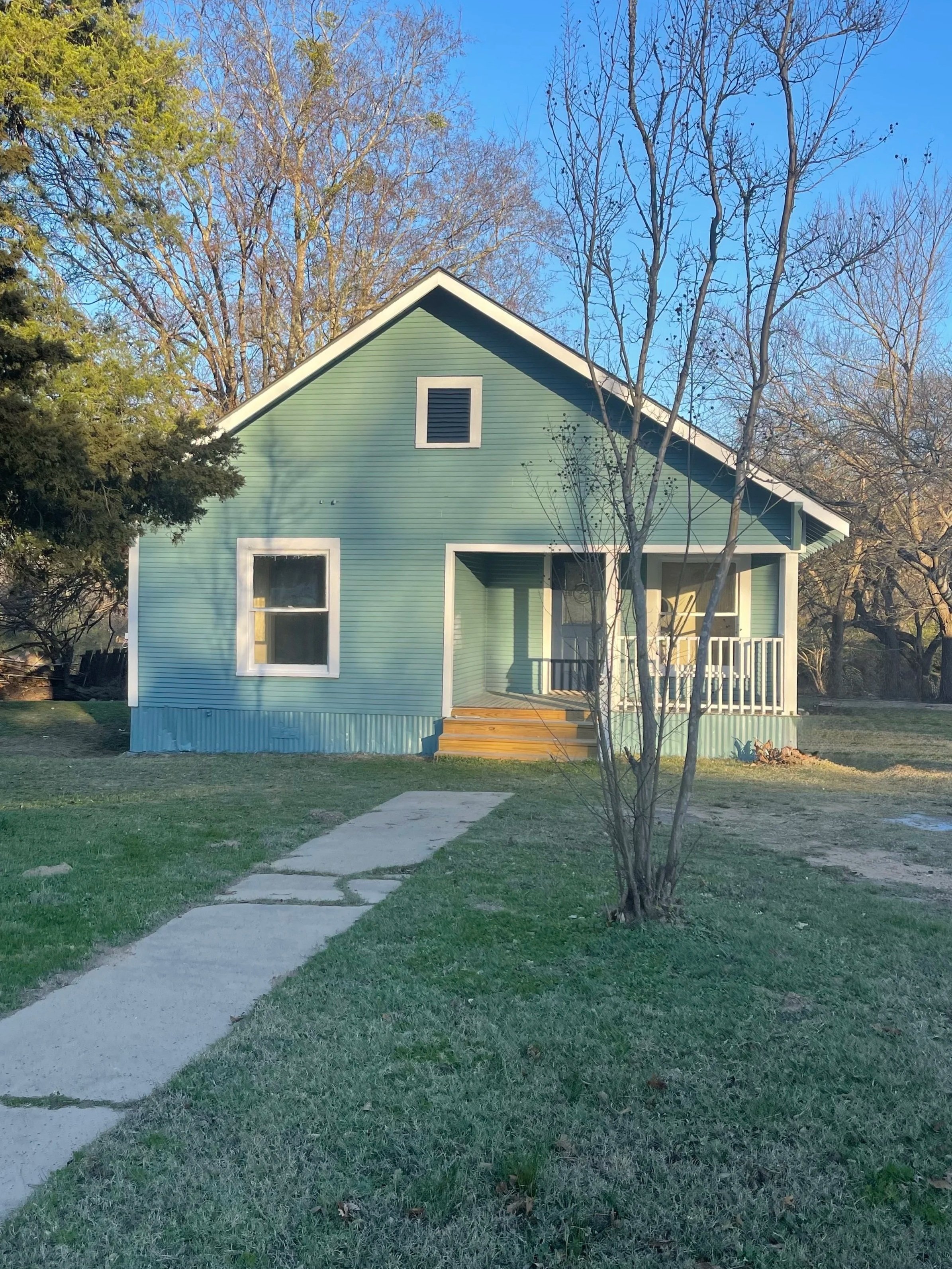A small green house with white trim and a front porch with wooden stairs. There is a concrete walkway leading up to the house, and a young leafless tree in the front yard. Tall trees surround the house, and it is sunny with a blue sky.