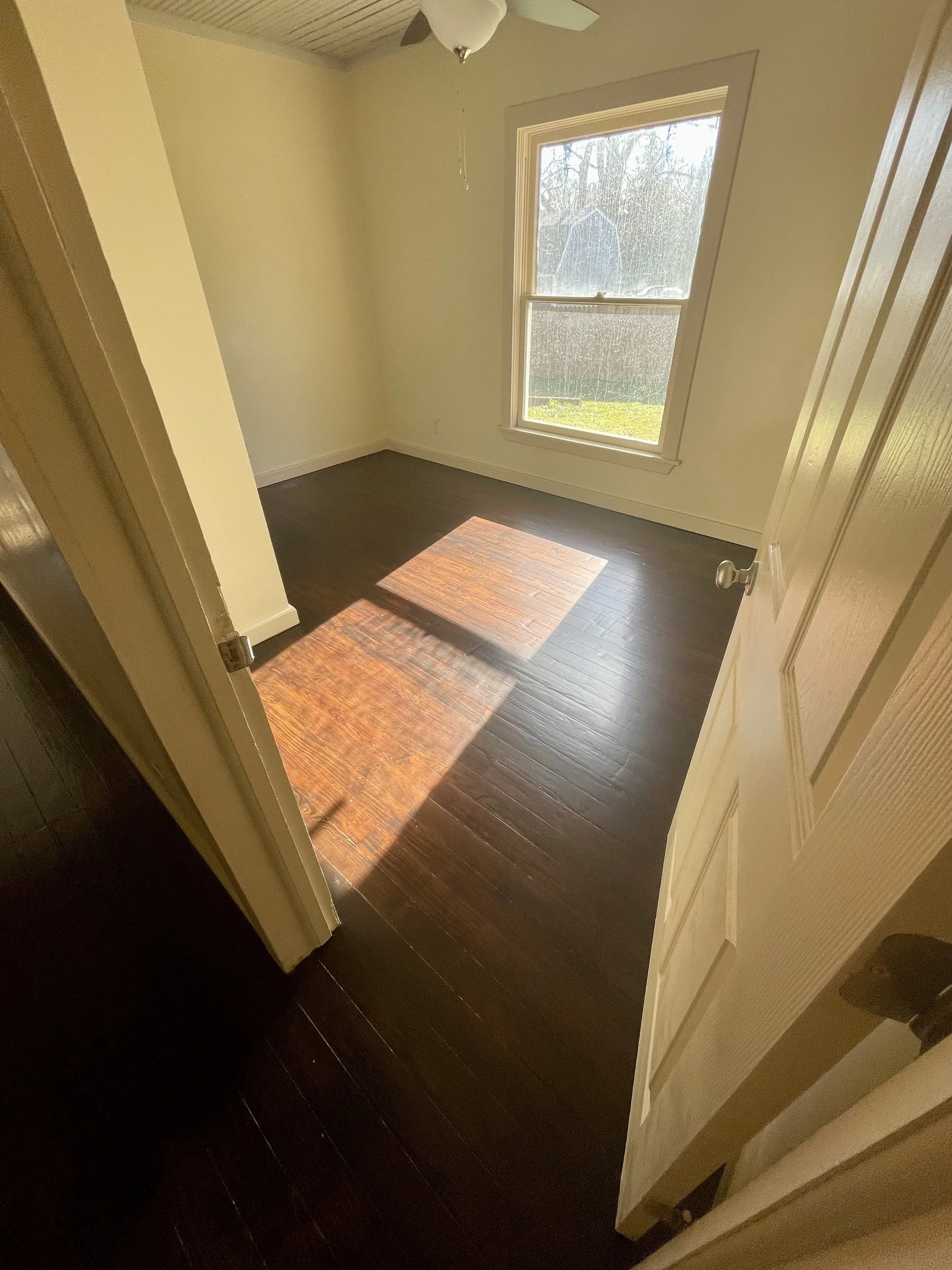 Empty room with hardwood floor, white walls, a large window letting in sunlight, and a ceiling fan.