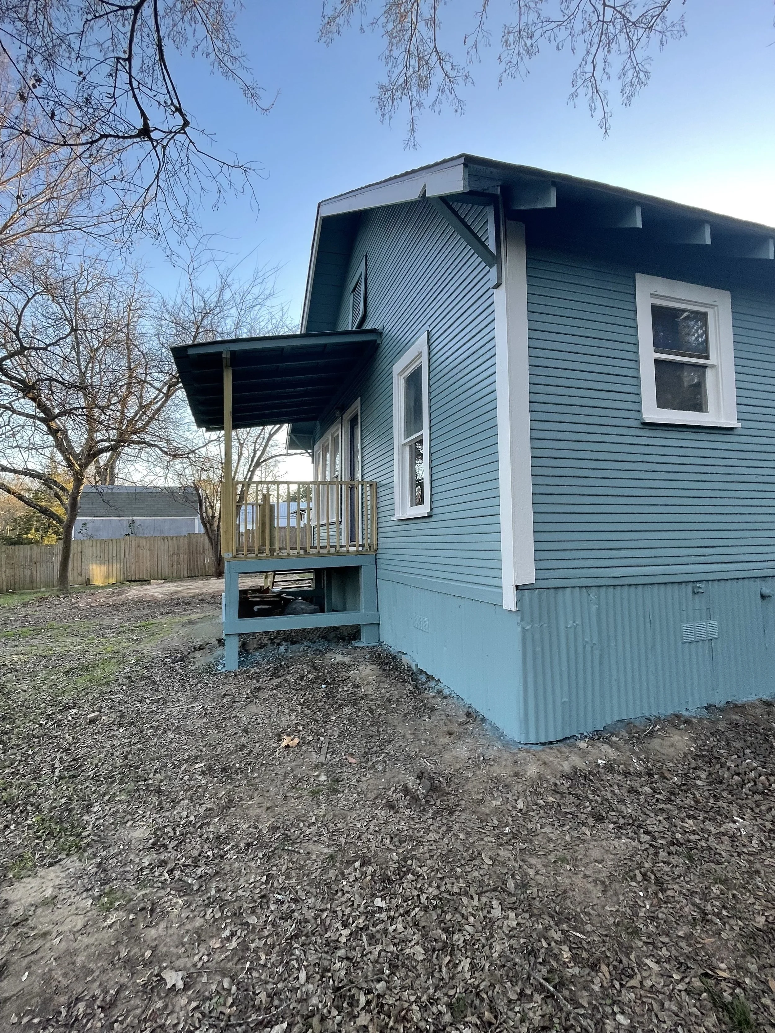 A house with blue siding, white trim, and a small wooden deck in the backyard, surrounded by a dirt and leaf-covered yard, with a few bare trees and a clear blue sky.