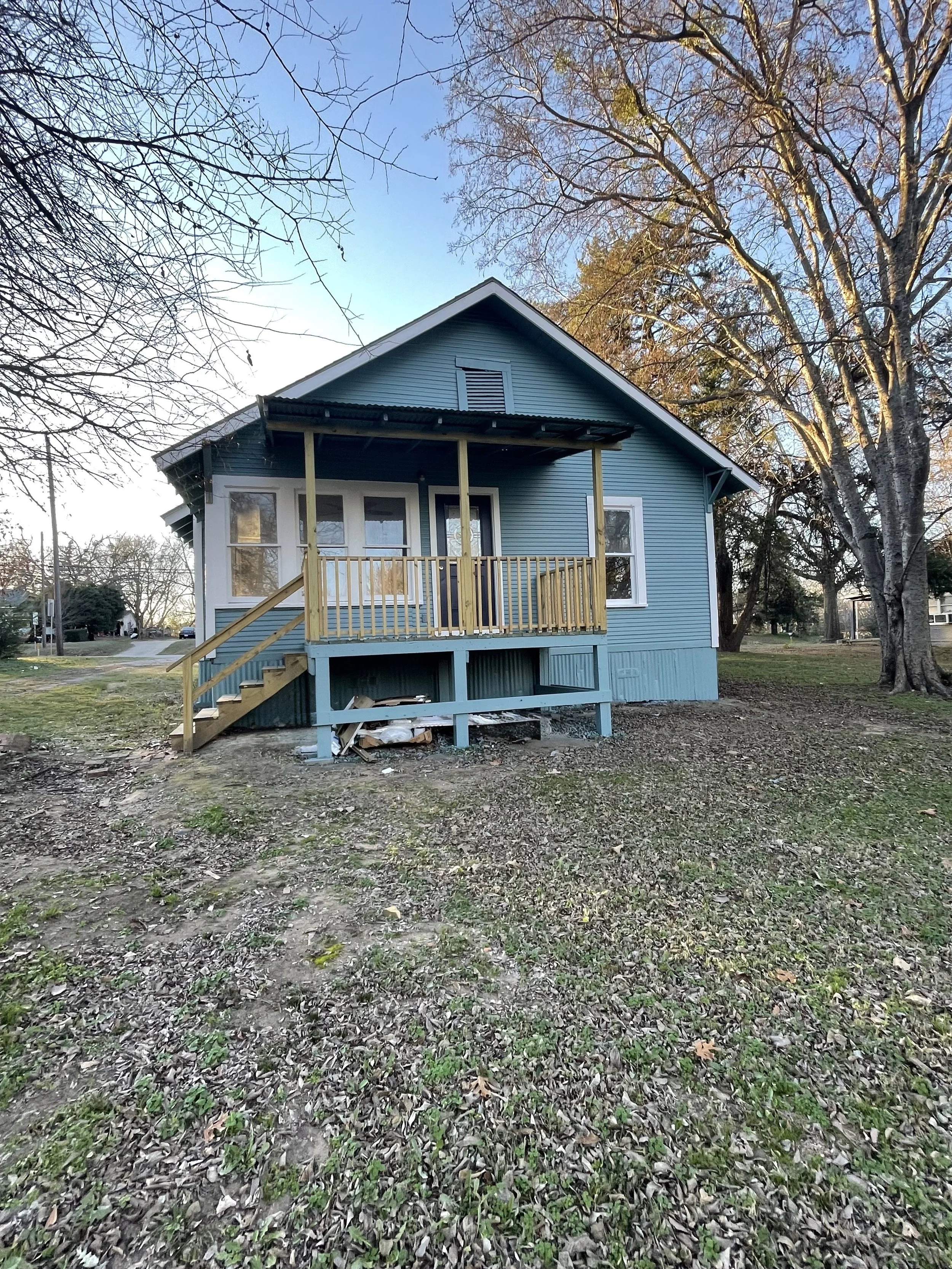 A house with blue weatherboard siding and a small front porch with wooden railings and steps, situated in a yard with sparse grass and patches of bare soil, and surrounded by bare trees under a clear sky.