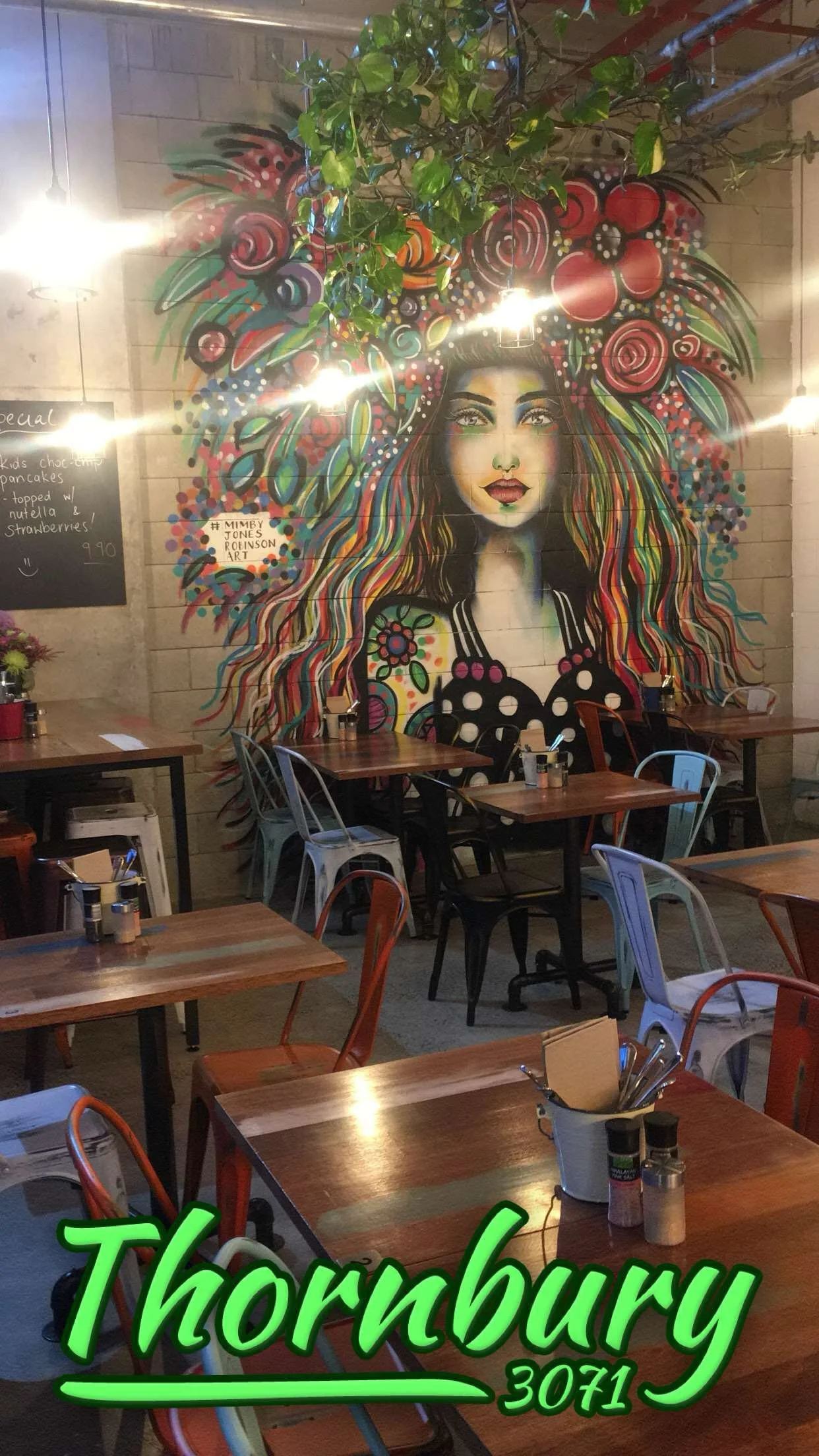 Colorful mural of a woman with long, multicolored hair and flowers in her hair on a wall inside a restaurant. Several tables and chairs are visible in the foreground.
